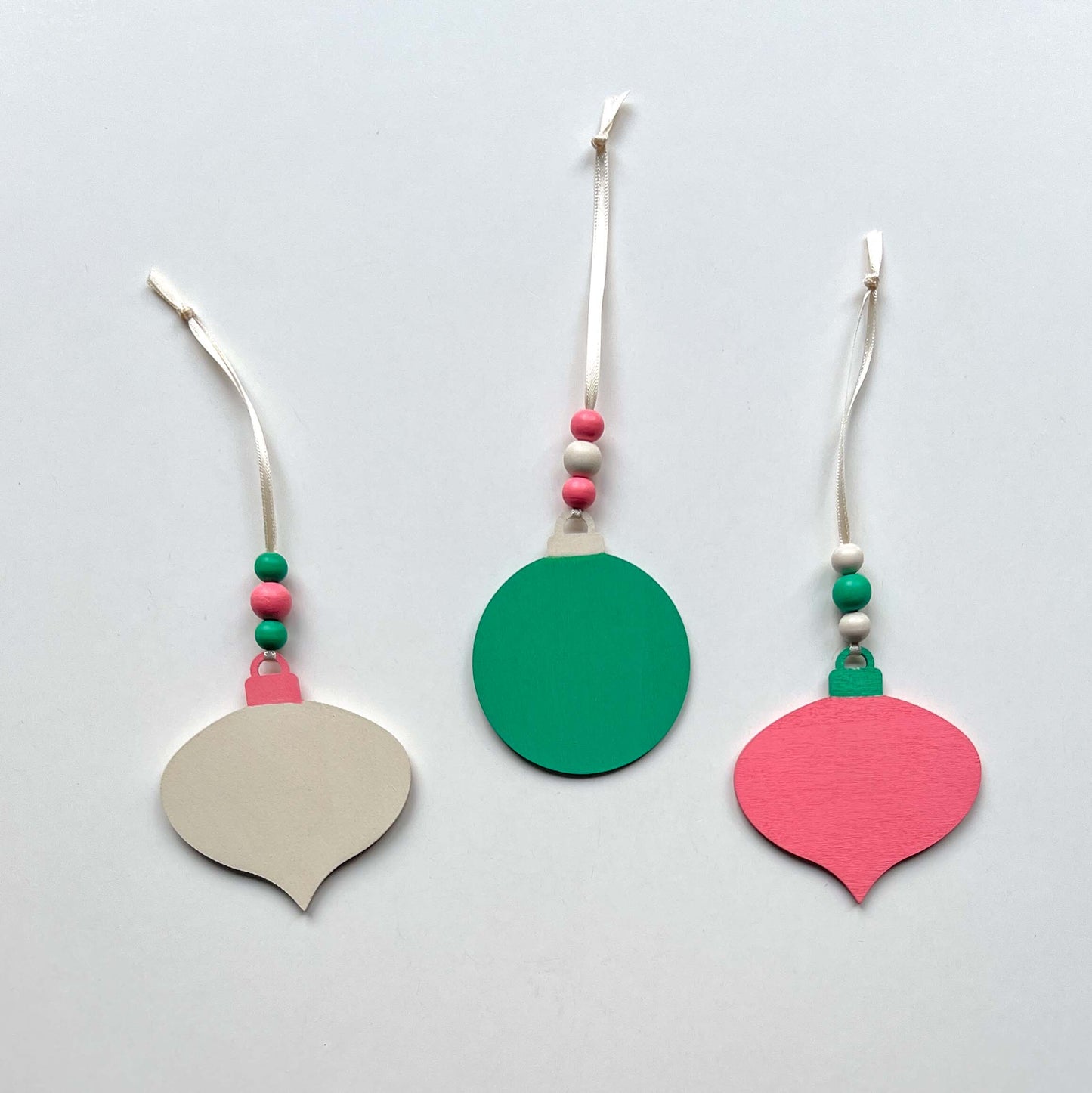 Back of retro themed wood Christmas ornaments painted bright pink, teal, and antique white. Each ornament hangs from a cream ribbon with three painted wood beads.