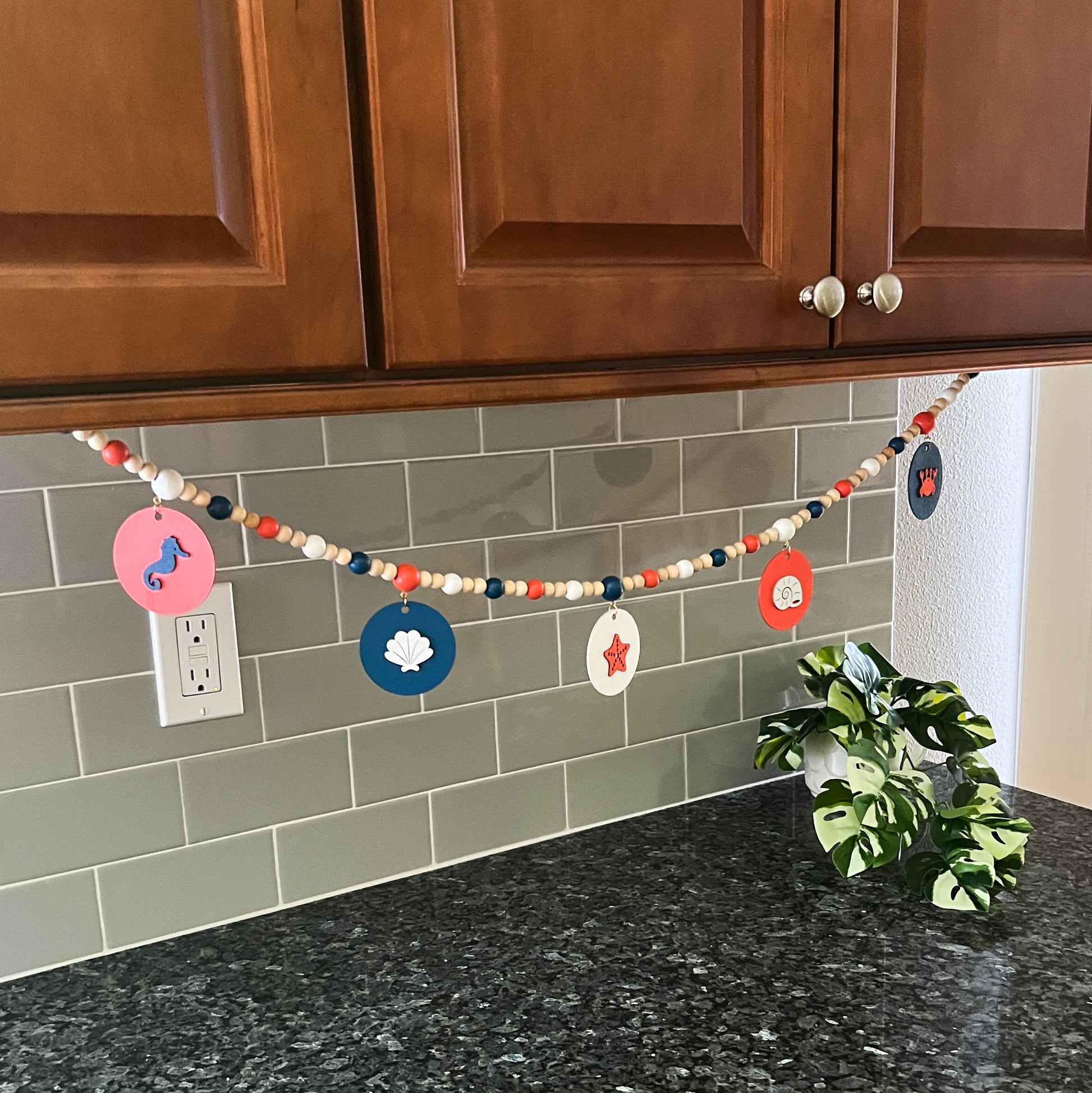 At the Seashore Handmade Garland. Features beachy wood shapes: seahorse, shells, starfish, and a crab. Shapes hang from painted wood beads. Summer beach seaside decor.