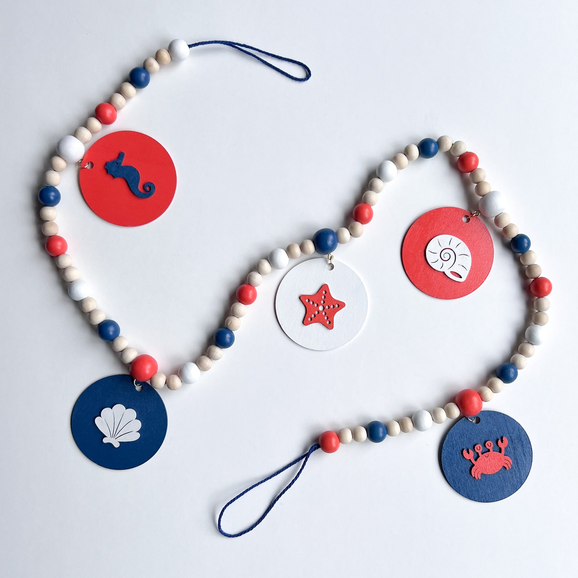At the Seashore Handmade Garland. Features beachy wood shapes: seahorse, shells, starfish, and a crab. Shapes hang from painted wood beads. Summer beach seaside decor.