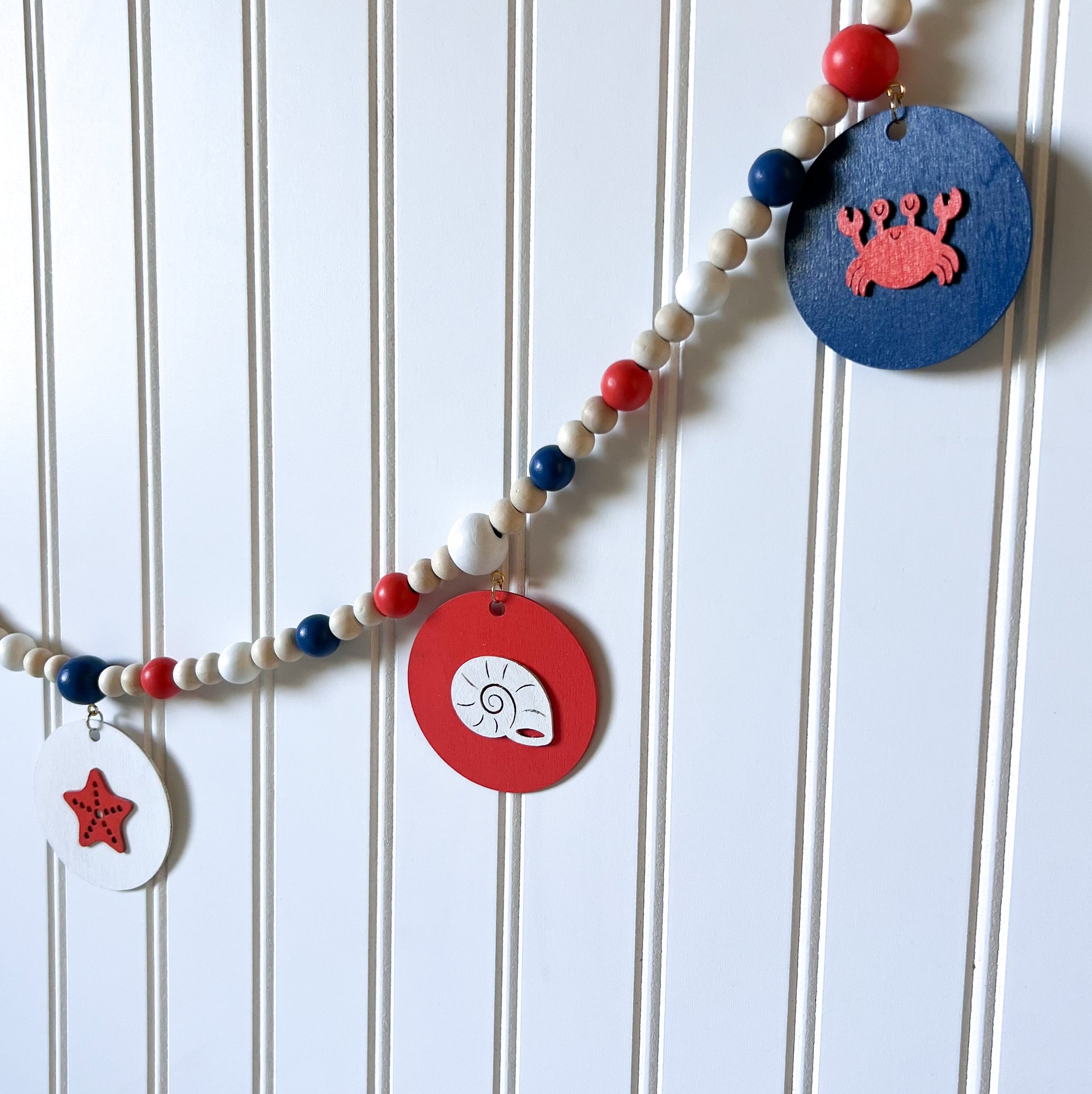 At the Seashore Handmade Garland. Features beachy wood shapes: seahorse, shells, starfish, and a crab. Shapes hang from painted wood beads. Summer beach seaside decor.