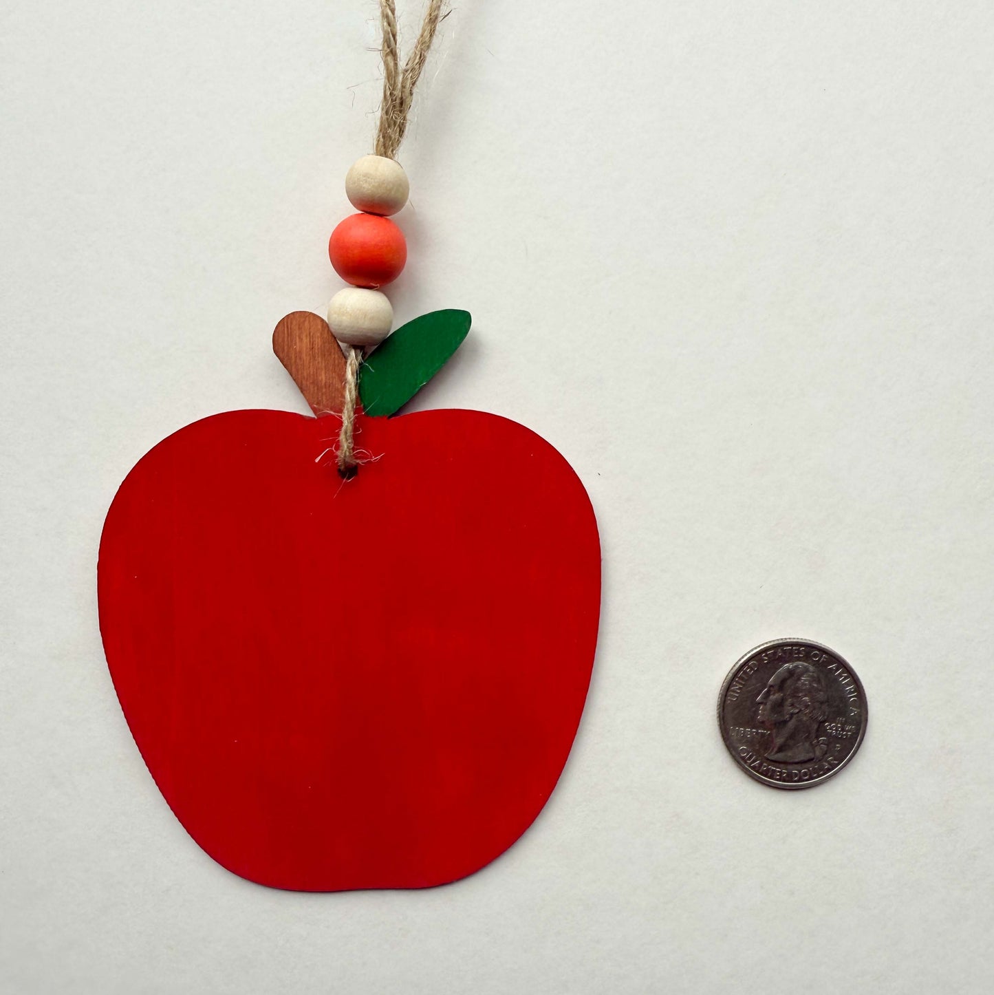 A wood apple ornament painted in bright red. Ornament hangs from jute twine and features three wood beads. Ornament is placed next to a quarter for size reference.