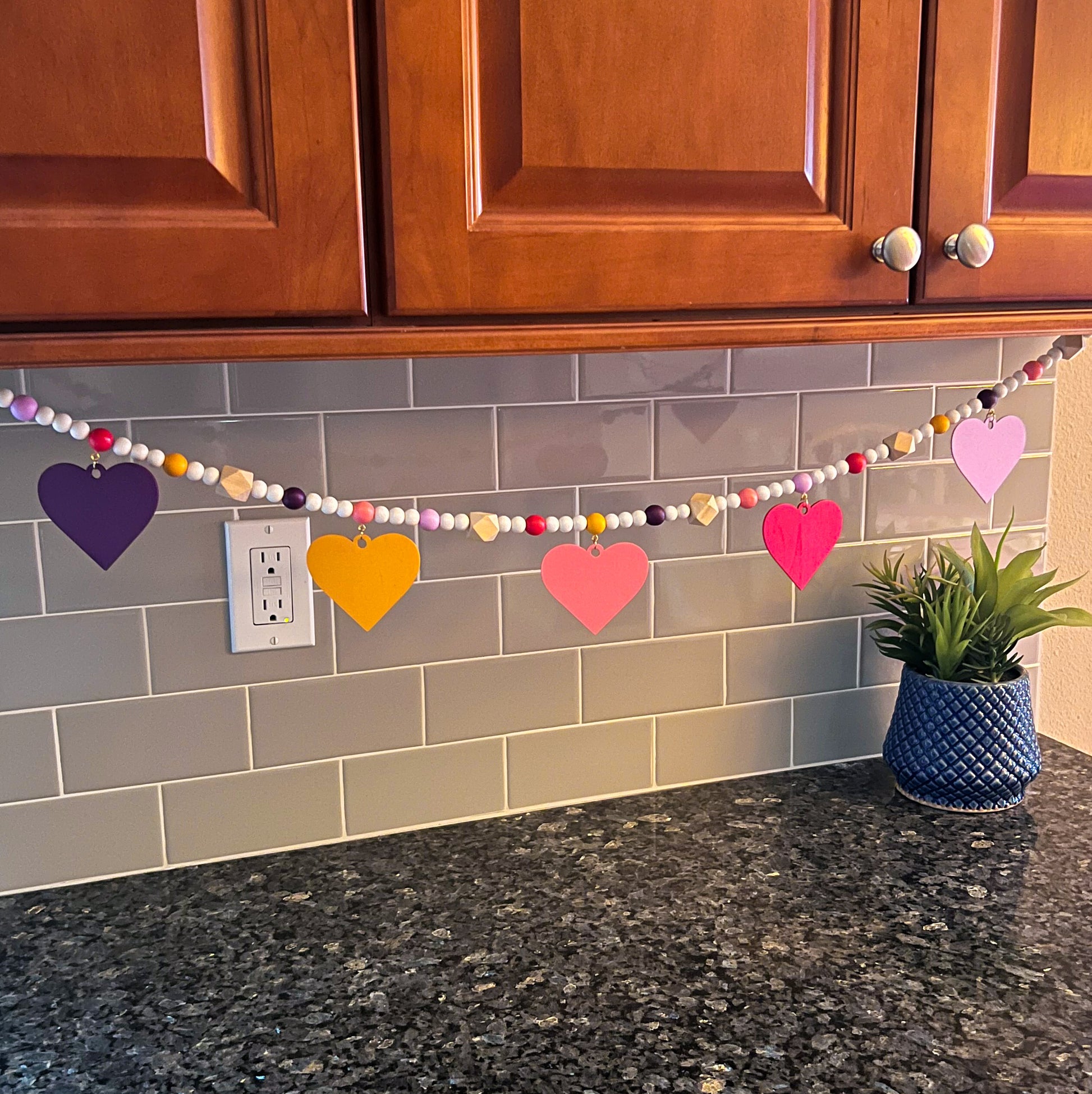 Images shows a garland with wood hearts in various colors – lavender, plum, golden yellow, bright pink, and pinkish-red. Hearts hang from natural geometric wood beads with a gold jump ring and eye screw. Those beads are strung with beads in corresponding colors to the hearts and smaller white beads on 2mm white macrame cord. 4-inch loops on each end make hanging the garland easy.