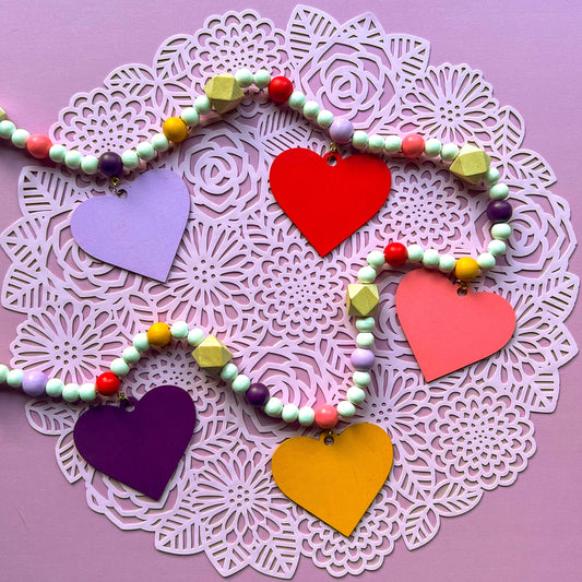 Images shows a garland with wood hearts in various colors – lavender, plum, golden yellow, bright pink, and pinkish-red. Hearts hang from natural geometric wood beads with a gold jump ring and eye screw. Those beads are strung with beads in corresponding colors to the hearts and smaller white beads on 2mm white macrame cord.