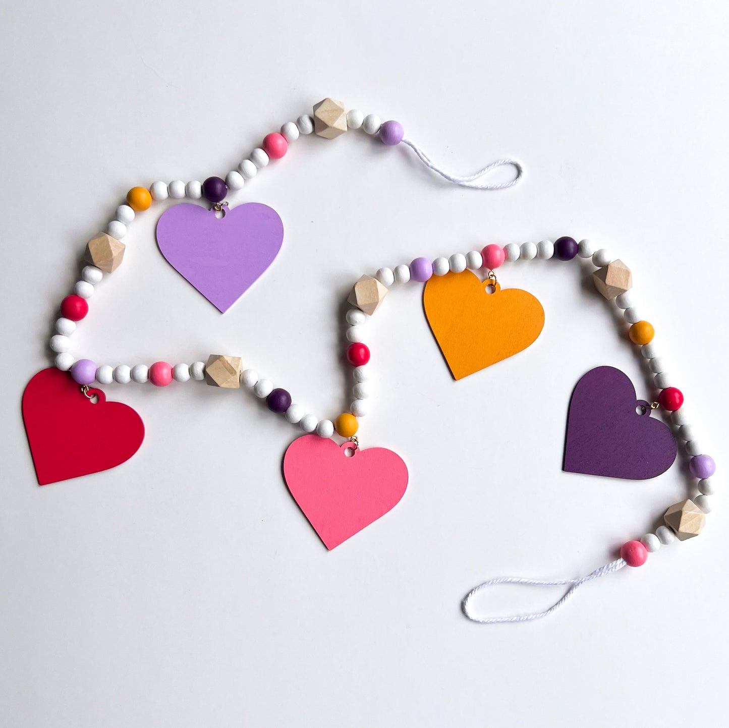 Images shows a garland with wood hearts in various colors – lavender, plum, golden yellow, bright pink, and pinkish-red. Hearts hang from natural geometric wood beads with a gold jump ring and eye screw. Those beads are strung with beads in corresponding colors to the hearts and smaller white beads on 2mm white macrame cord. 4-inch loops on each end make hanging the garland easy.