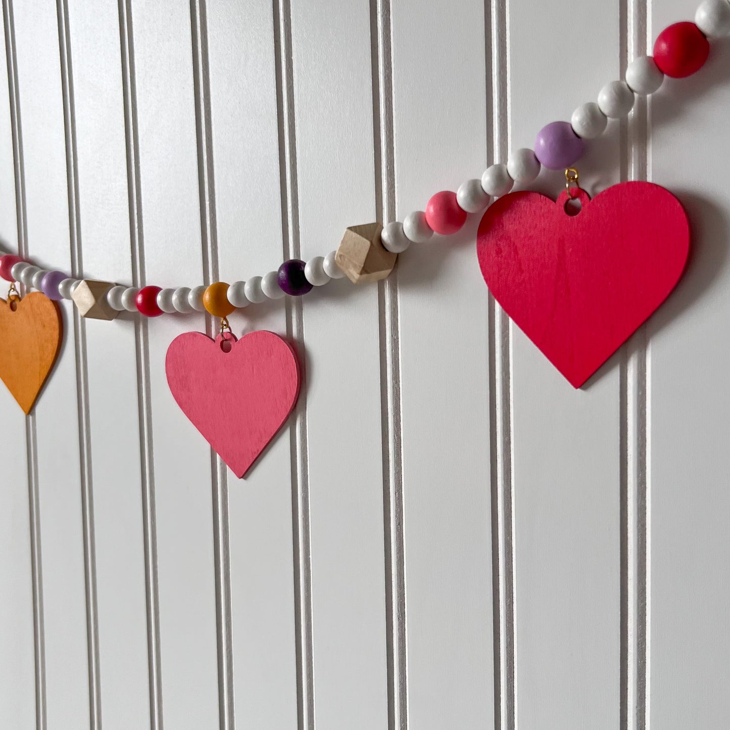 Images shows a garland with wood hearts in various colors – lavender, plum, golden yellow, bright pink, and pinkish-red. Hearts hang from natural geometric wood beads with a gold jump ring and eye screw. Those beads are strung with beads in corresponding colors to the hearts and smaller white beads on 2mm white macrame cord. 4-inch loops on each end make hanging the garland easy.