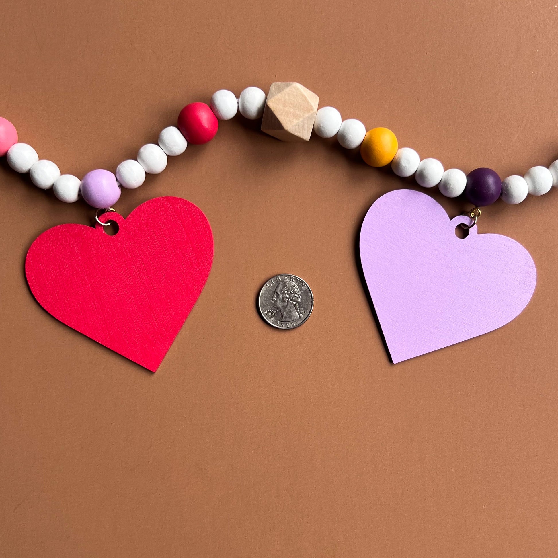 Images shows a garland with wood hearts in various colors – lavender, plum, golden yellow, bright pink, and pinkish-red. Hearts hang from natural geometric wood beads with a gold jump ring and eye screw. Those beads are strung with beads in corresponding colors to the hearts and smaller white beads on 2mm white macrame cord. Garland is placed next to a quarter as a size comparison.