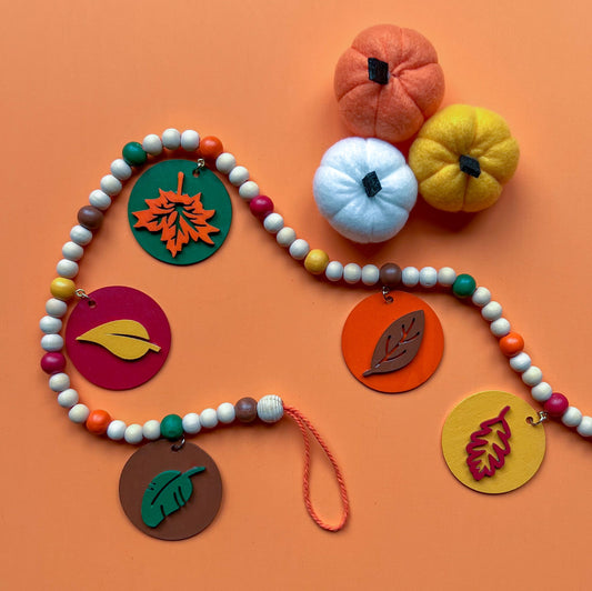 Image shows a wood bead garland with 2" wood circles that feature various colored fall leaves. Circles hang from painted beads with natural wood beads are interspersed. Beads are strung on 2mm orange macrame cord with 3" loops on each end.