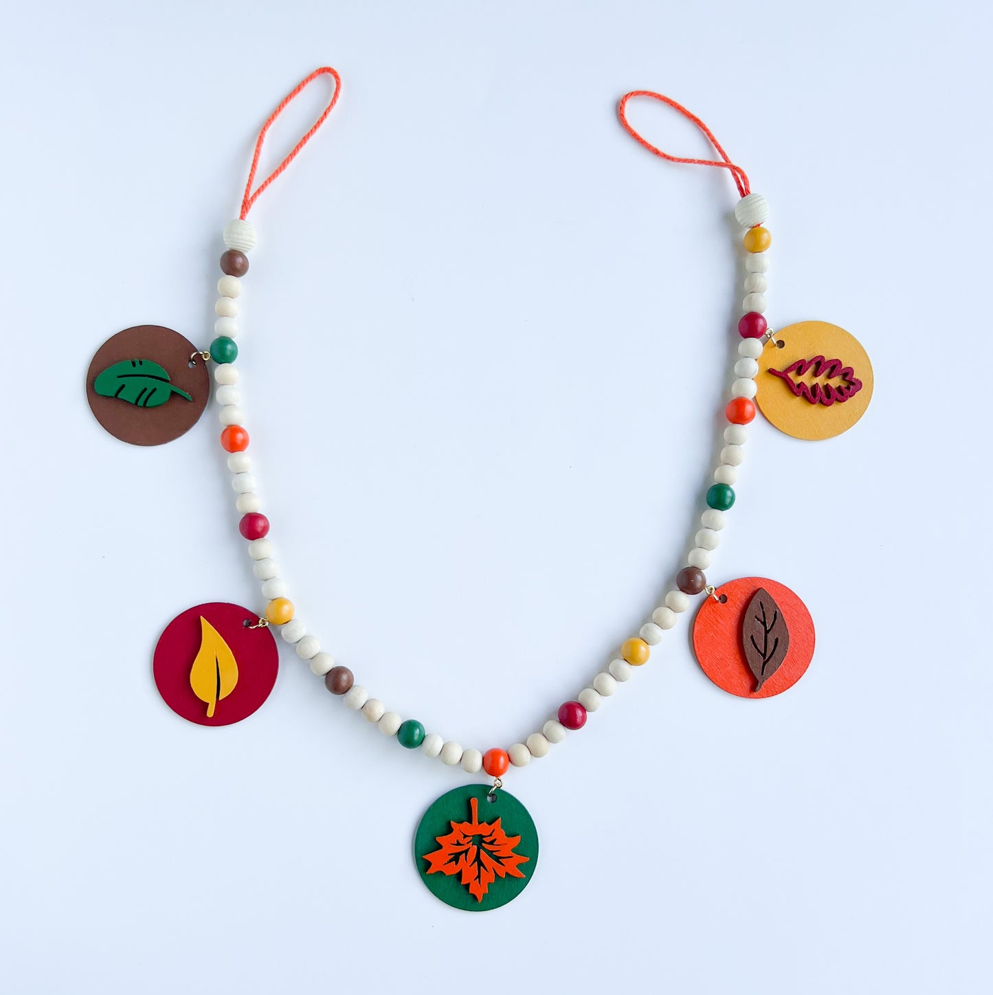 Image shows a wood bead garland with 2" wood circles that feature various colored fall leaves. Circles hang from painted beads with natural wood beads are interspersed. Beads are strung on 2mm orange macrame cord with 3" loops on each end.