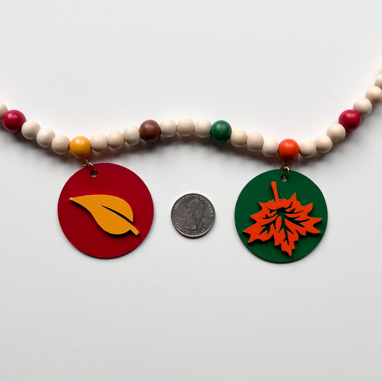 Image shows a closeup of a wood bead garland with 2" wood circles that feature various colored fall leaves. Circles hang from painted beads with natural wood beads are interspersed. Beads are strung on 2mm orange macrame cord with 3" loops on each end.