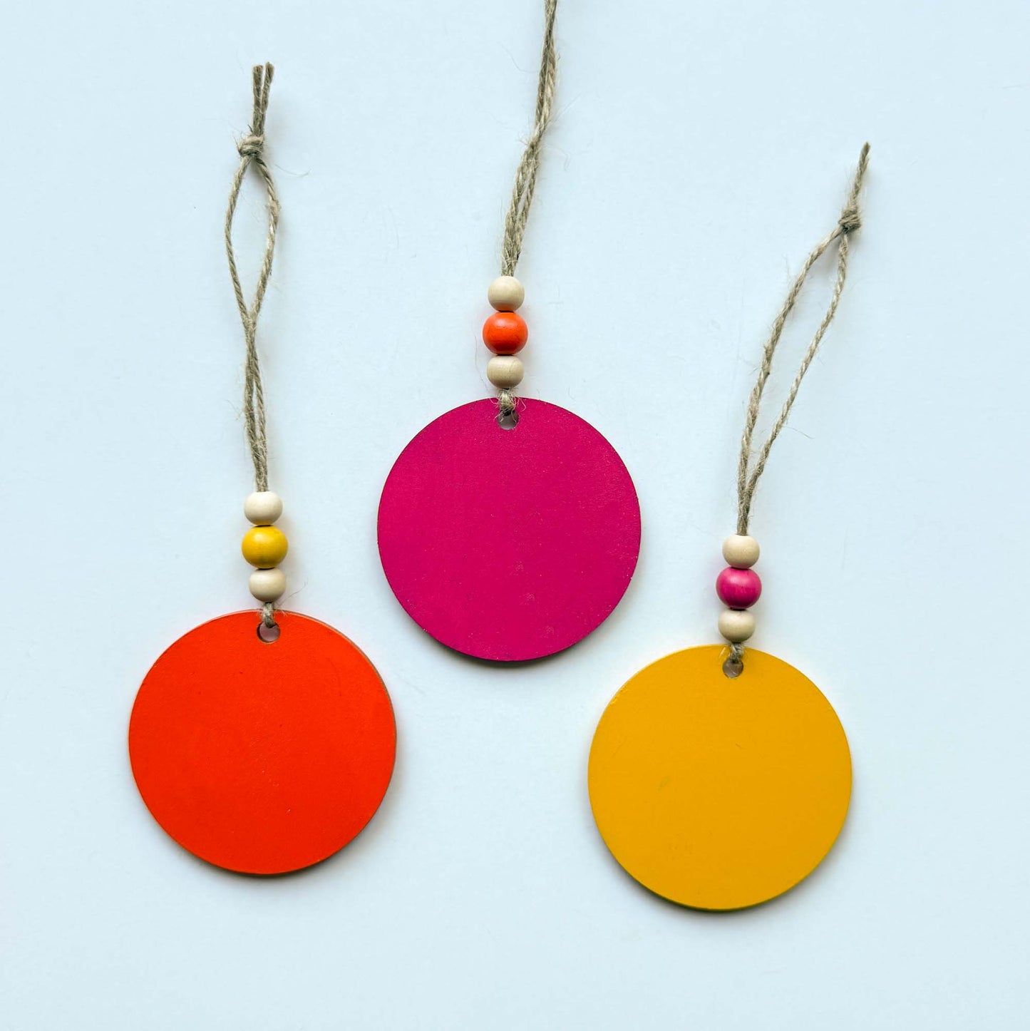 Back of set of 3 wood, tropical themed ornaments. Ornaments are flat wood circles measuring 2.75 inches; each features a different monstera or palm leaf. Ornaments are painted in magenta, yellow, or orange and include three wood beads strung on jute twine.