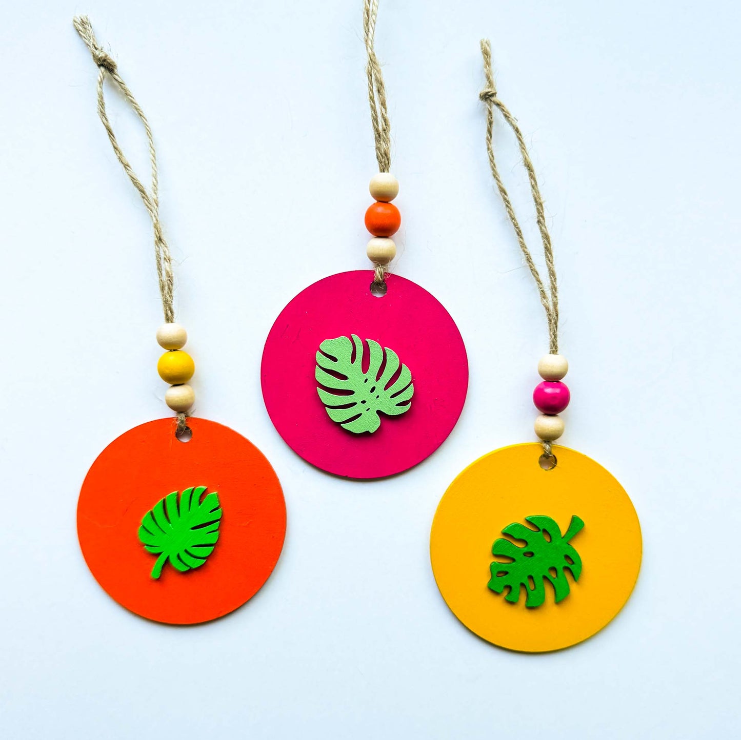Set of 3 wood, tropical themed ornaments. Ornaments are flat wood circles measuring 2.75 inches; each features a different monstera or palm leaf. Ornaments are painted in magenta, yellow, or orange and include three wood beads strung on jute twine.