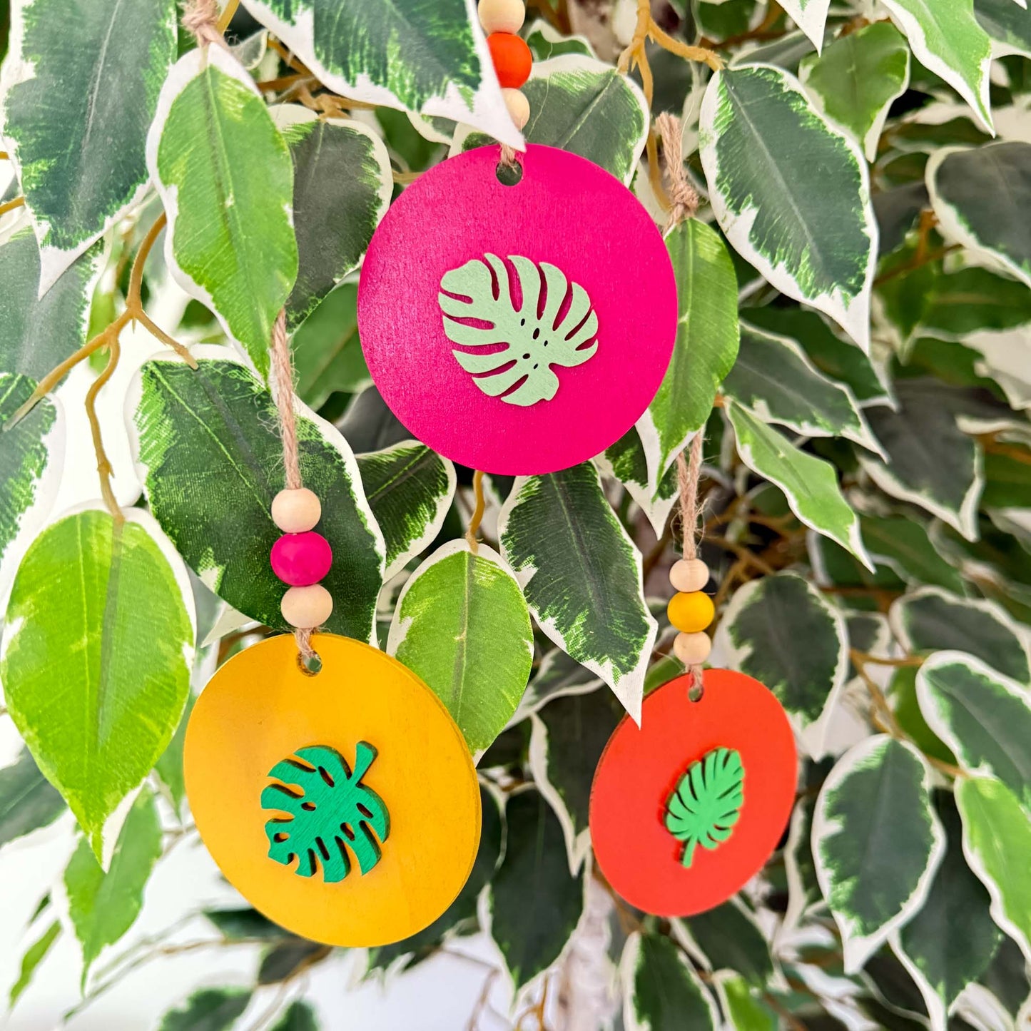 Set of 3 wood, tropical themed ornaments. Ornaments are flat wood circles measuring 2.75 inches; each features a different monstera or palm leaf. Ornaments are painted in magenta, yellow, or orange and include three wood beads strung on jute twine.