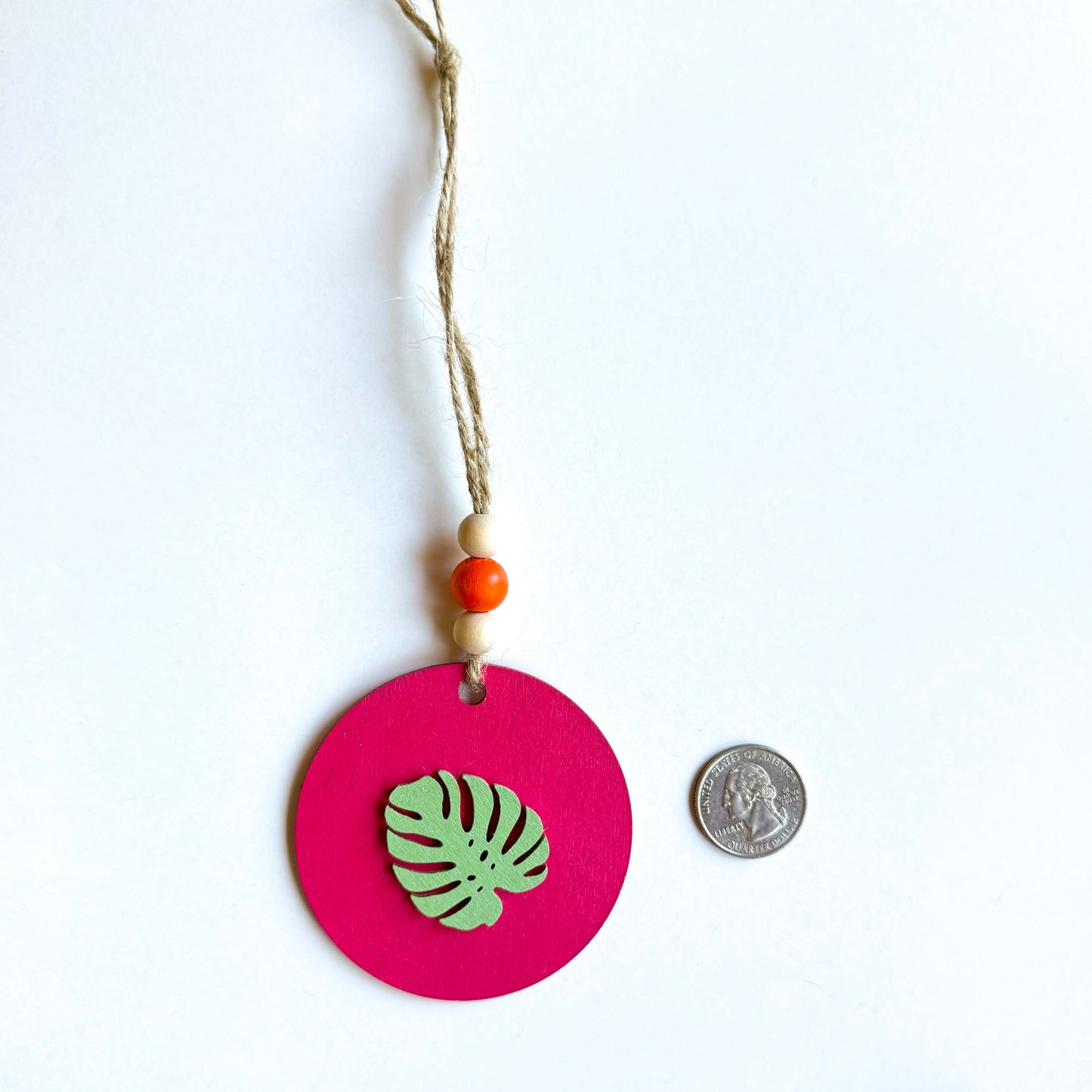 Tropical themed ornament featuring a 2.75 inch flat wood circle painted in magenta with a green monstera leaf. Ornament hangs from jute twine with three wood beads. A quarter is placed next to the ornament as a size comparison.