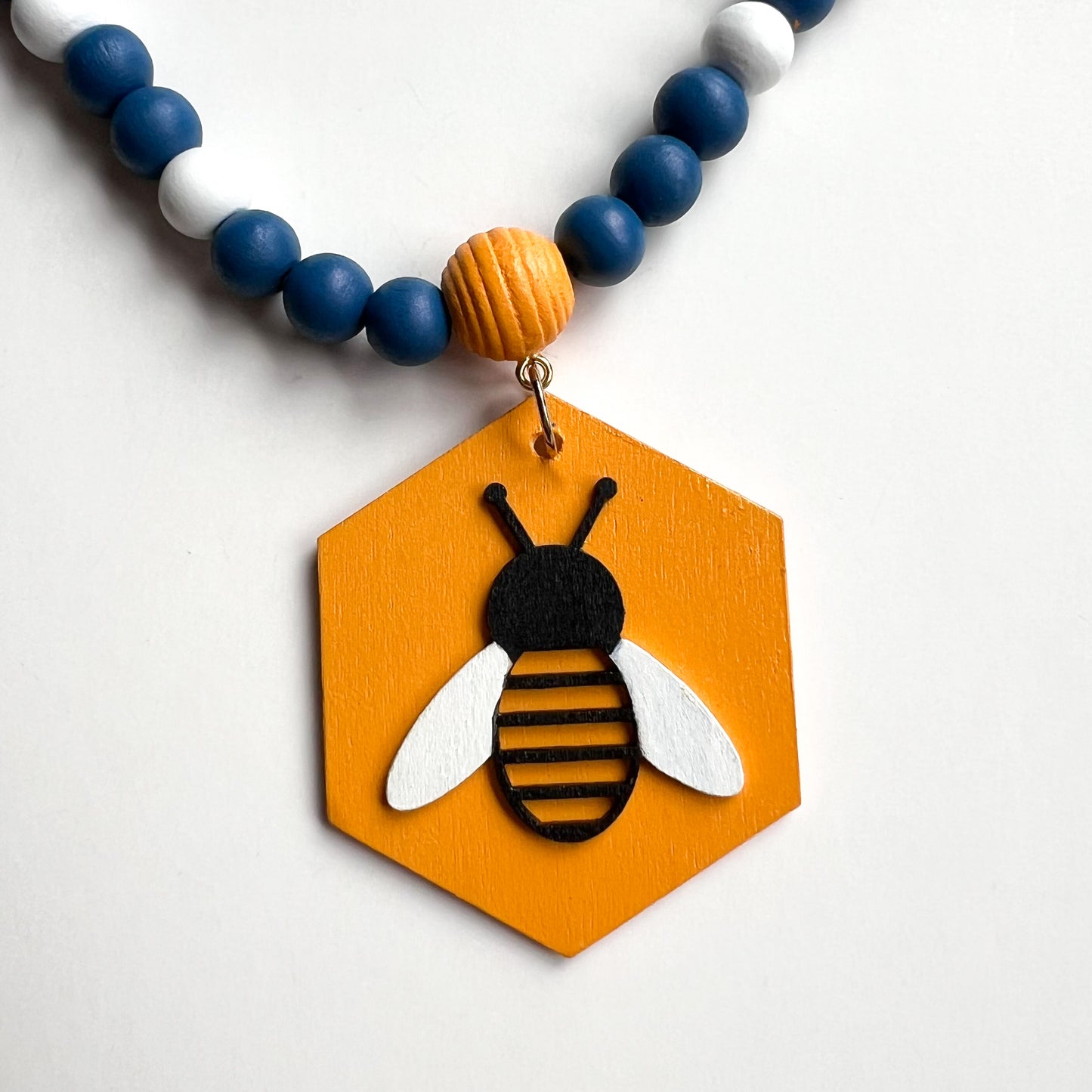 The image shows a closeup of a wood bead garland featuring wood hexagons with bees and flowers, strung with beads painted deep blue, golden yellow, and white. Hexagons hang from beehive shaped beads. Beads are strung on 2mm blue macrame cord with 3-inch loops on each end.