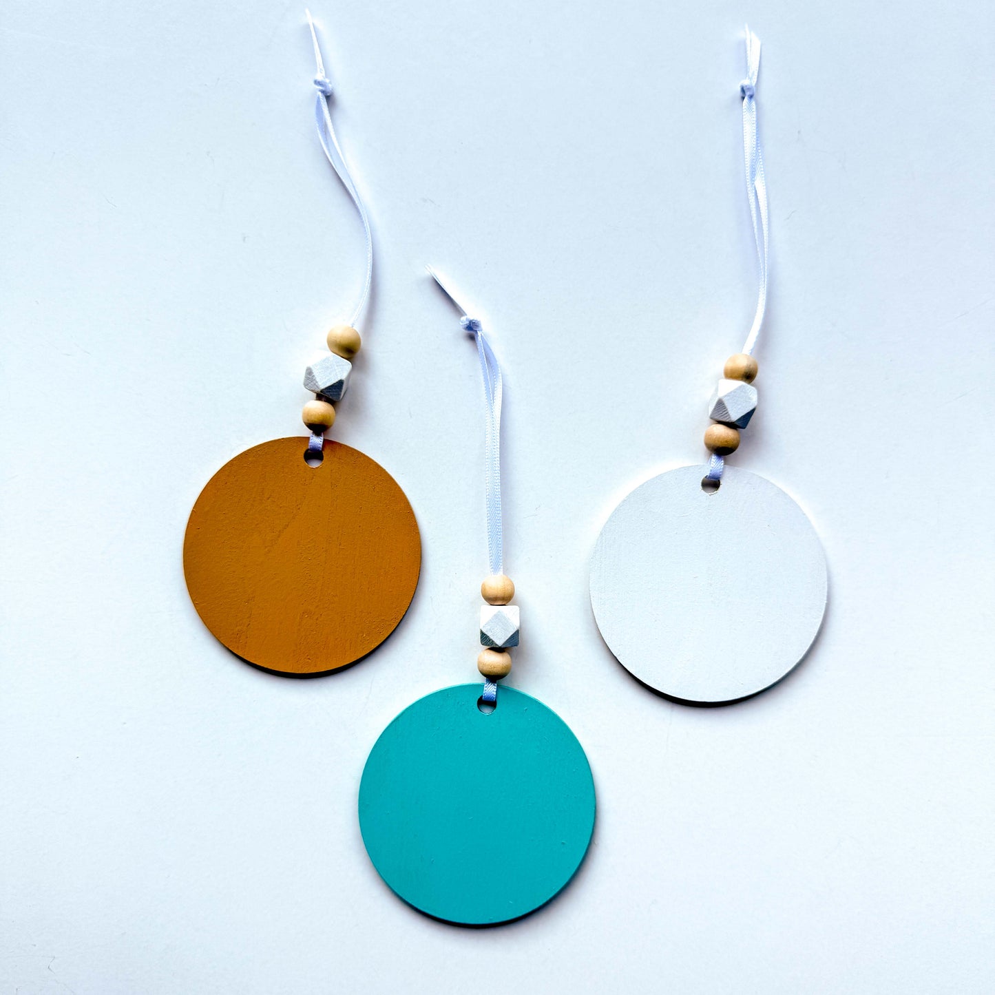 The back of a set of three coastal themed ornaments on flat wood rounds in colors of bright blue, white, and sand. A coast themed shape in on each ornament, including a starfish and two different shell shapes. Ornaments hang from a white ribbon and feature two natural colored beads and a white geometric bead as an accent.