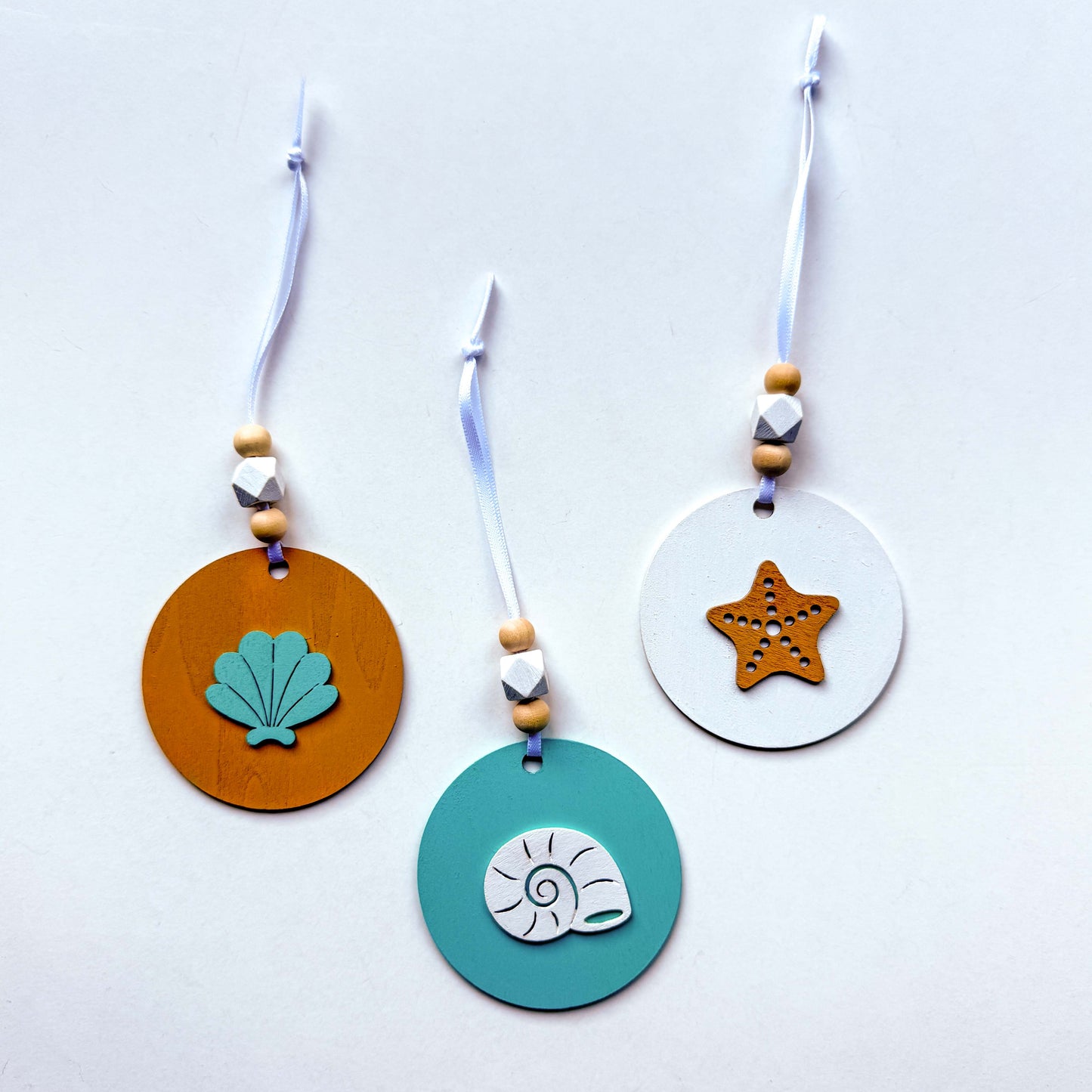 Set of three coastal themed ornaments on flat wood rounds in colors of bright blue, white, and sand. A coast themed shape in on each ornament, including a starfish and two different shell shapes. Ornaments hang from a white ribbon and feature two natural colored beads and a white geometric bead as an accent.