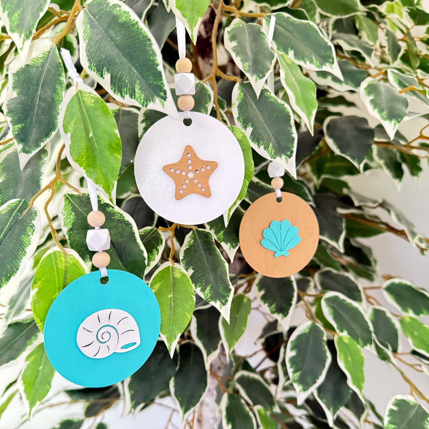 Set of three coastal themed ornaments on flat wood rounds in colors of bright blue, white, and sand. A coast themed shape in on each ornament, including a starfish and two different shell shapes. Ornaments hang from a white ribbon and feature two natural colored beads and a white geometric bead as an accent.