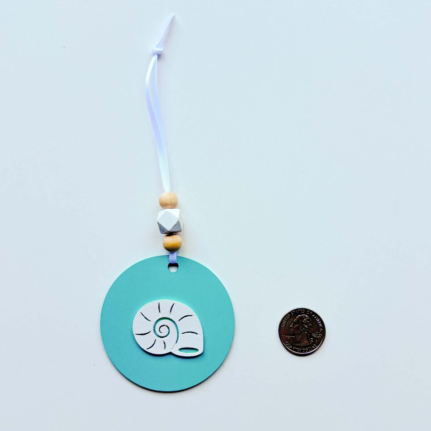 A coastal themed ornaments on flat wood round painted a bright blue featuring a white shell shape. Ornament hangs from a white ribbon and features two natural colored beads and a white geometric bead. A quarter sits next to the ornament as a size comparison.