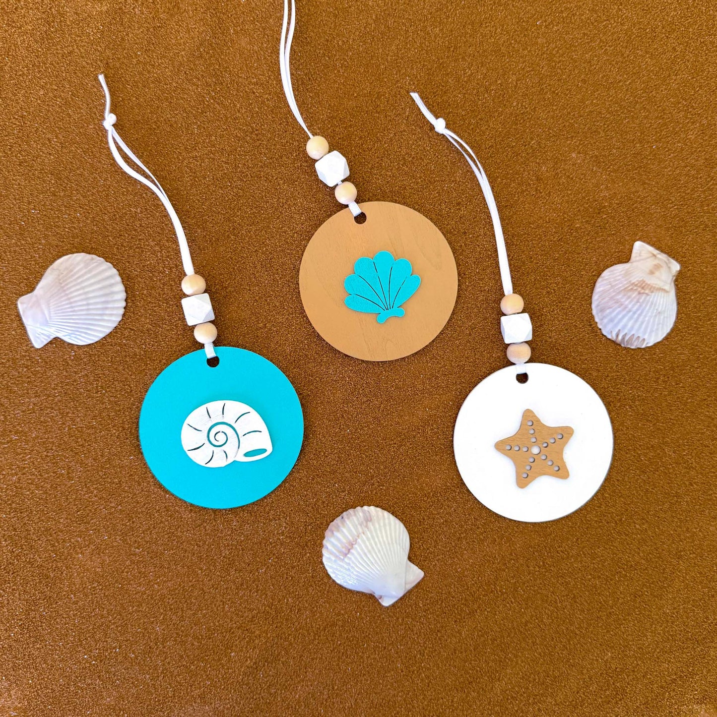 Set of three coastal themed ornaments on flat wood rounds in colors of bright blue, white, and sand. A coast themed shape in on each ornament, including a starfish and two different shell shapes. Ornaments hang from a white ribbon and feature two natural colored beads and a white geometric bead as an accent.