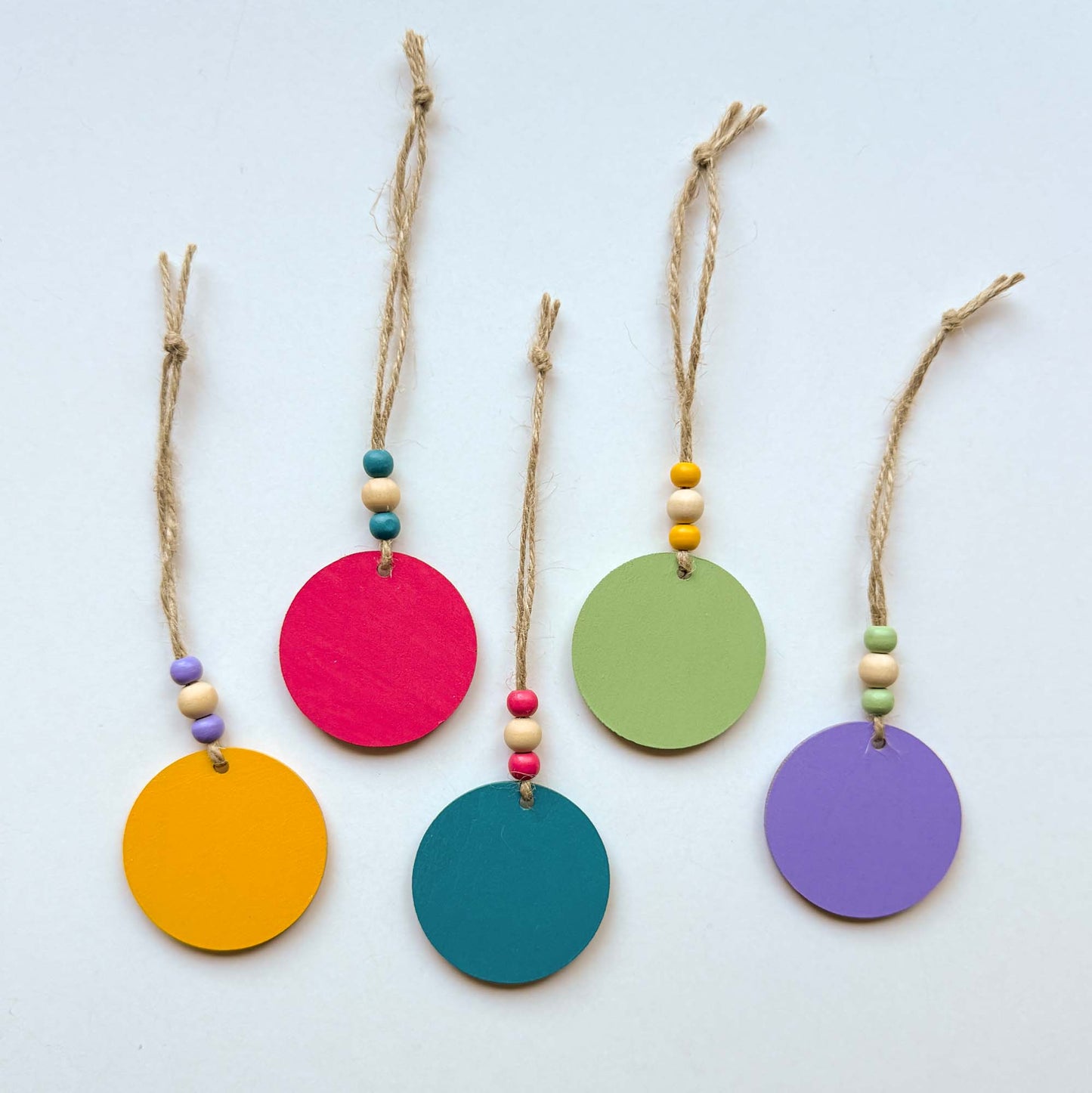 Back of small 2 inch round wood ornaments with tropical themed shapes including a pineapple, a cactus, a tropical leaf, a flower, and a bird. Ornaments hang from jute cording with three small beads. Painted in bright colors of magenta, yellow, lavender, green, and medium blue.