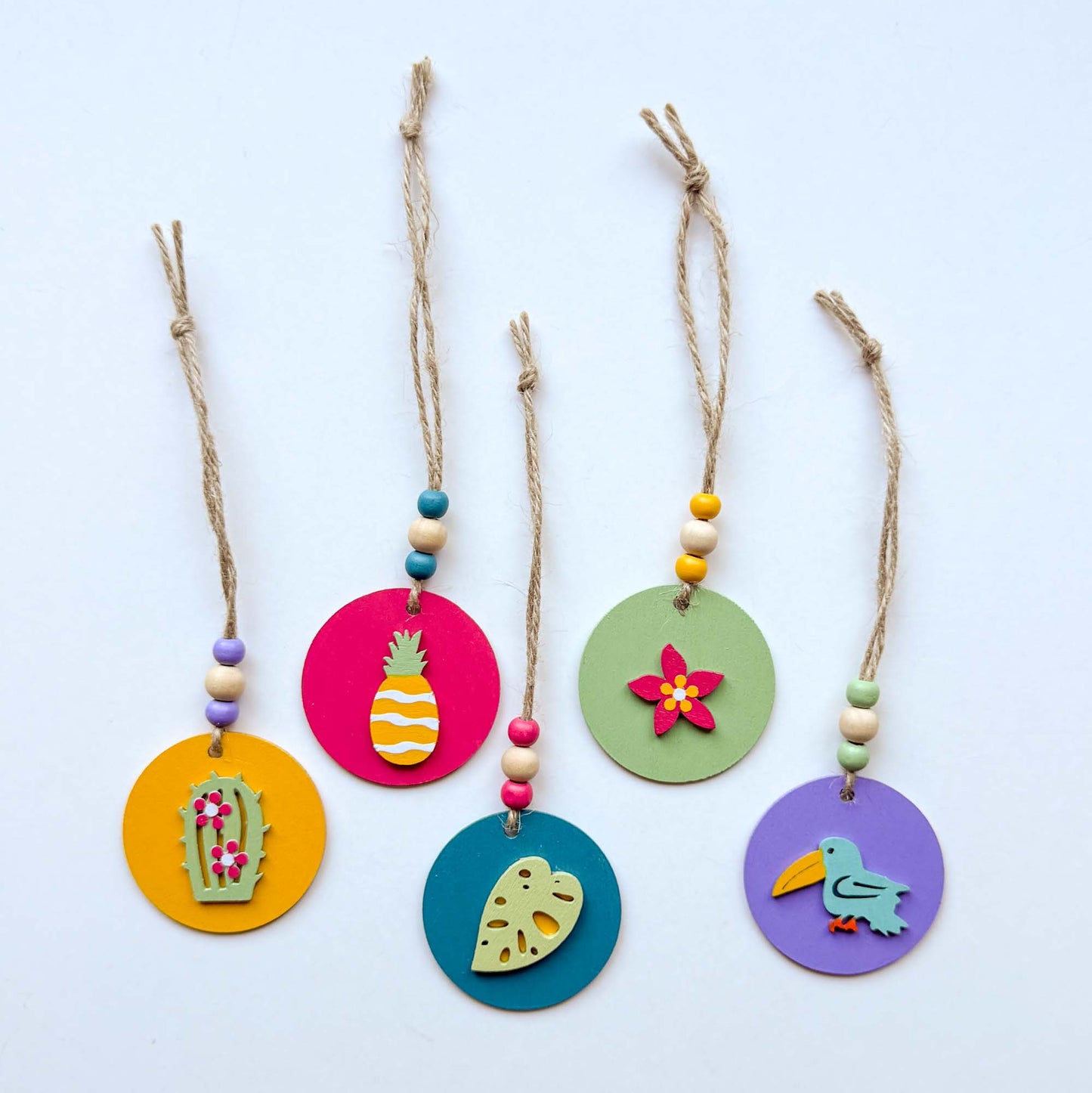 Small 2 inch round wood ornaments with tropical themed shapes including a pineapple, a cactus, a tropical leaf, a flower, and a bird. Ornaments hang from jute cording with three small beads. Painted in bright colors of magenta, yellow, lavender, green, and medium blue.