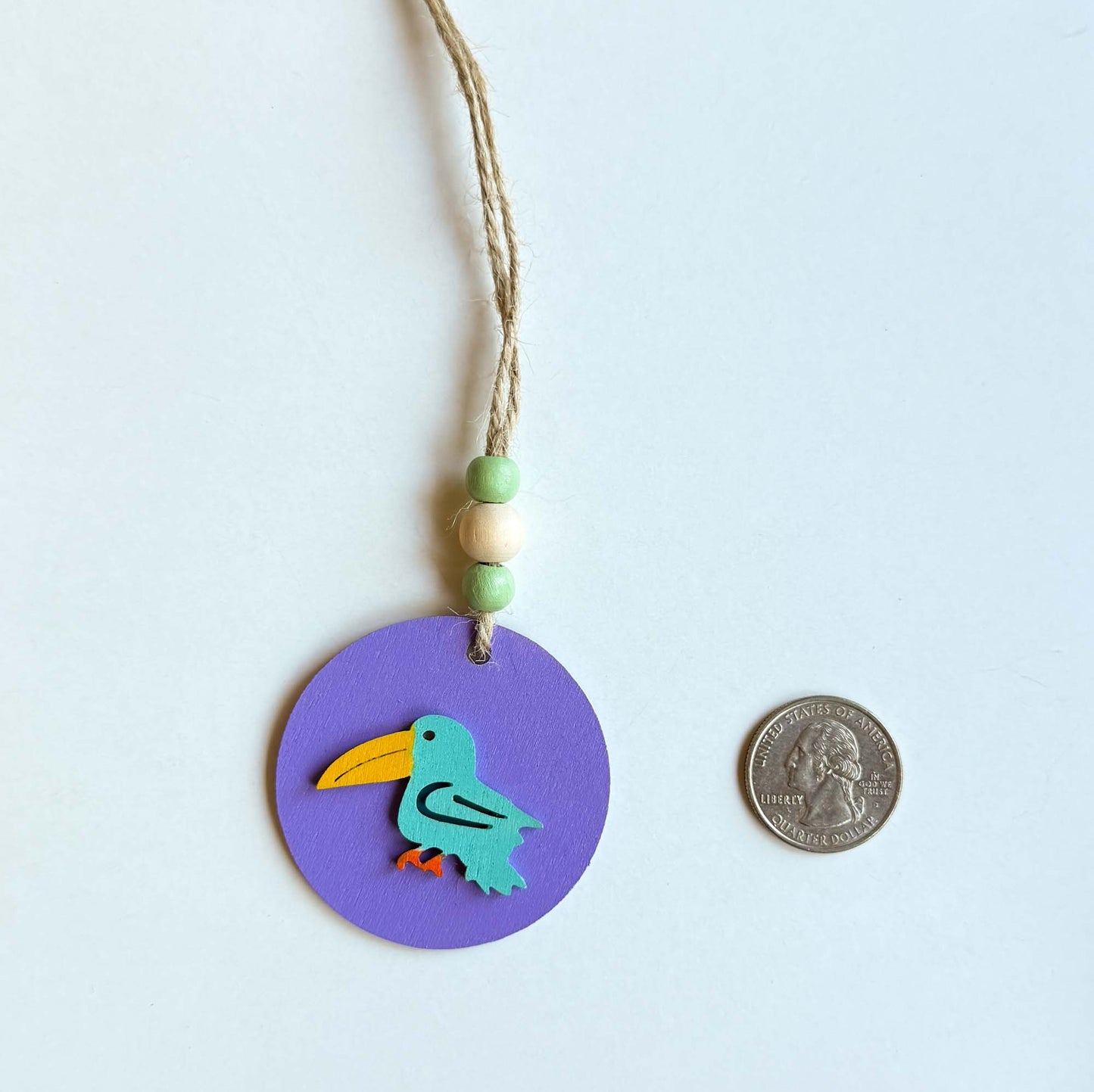 Small 2 inch round lavender wood ornament with tropical bird painted blue. Ornament hangs from jute cording with three small beads. A quarter is placed next to it for size comparison.