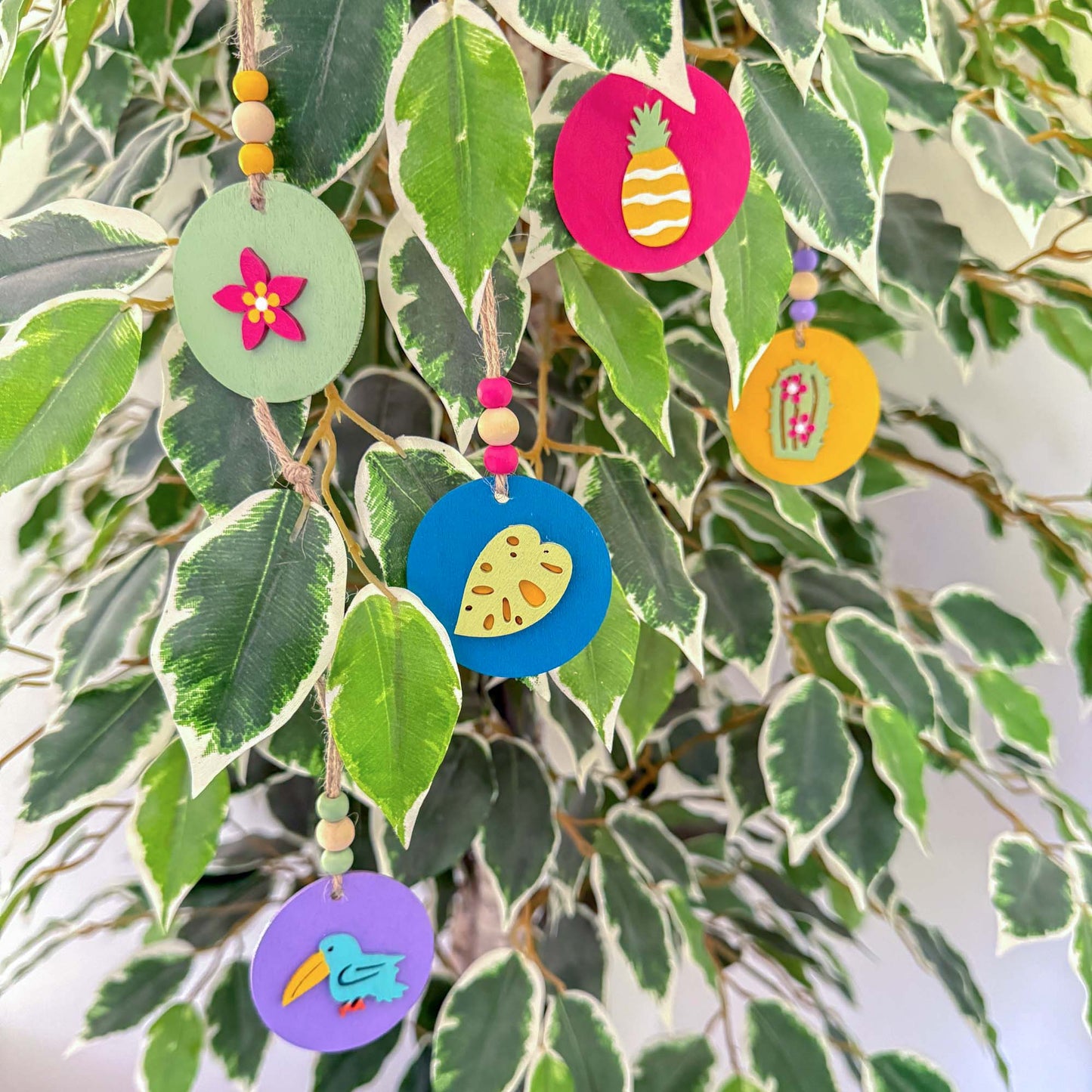 Small 2 inch round wood ornaments with tropical themed shapes including a pineapple, a cactus, a tropical leaf, a flower, and a bird. Ornaments hang from jute cording with three small beads. Painted in bright colors of magenta, yellow, lavender, green, and medium blue.