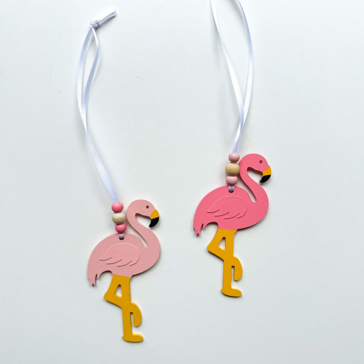 Two wood flamingo ornaments in different shades of pink hang from a 6" white ribbon with three beads in white and pink. Ornaments are on a white background.