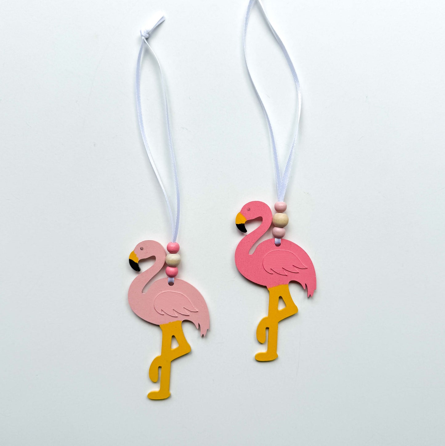 Two wood flamingo ornaments in different shades of pink hang from a 6" white ribbon with three beads in white and pink. Ornaments are on a white background.