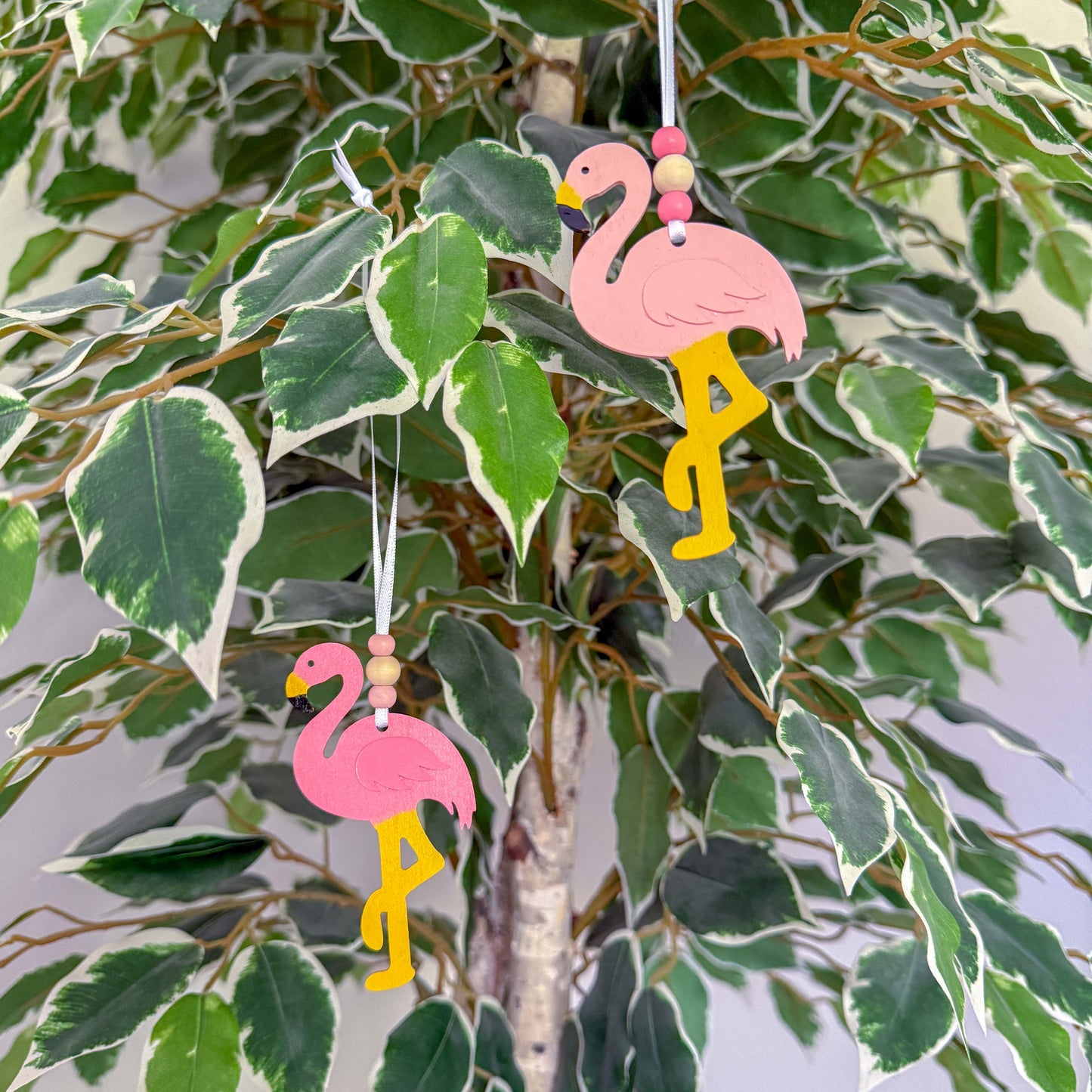 Two wood flamingo ornaments in different shades of pink hang from a 6" white ribbon with three beads in white and pink. Ornaments hang from a seasonal tree branch.