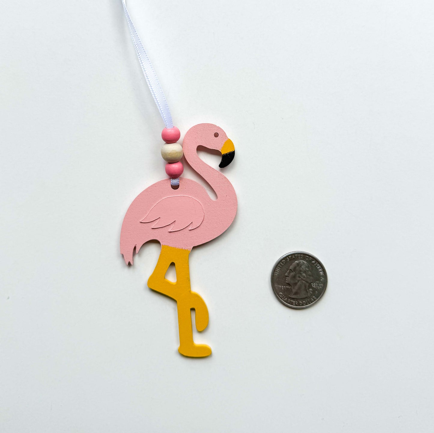 Wood flamingo ornaments in light pink that hangs from a 6" white ribbon with three beads in white and pink. Ornaments is placed next to a quarter for reference.
