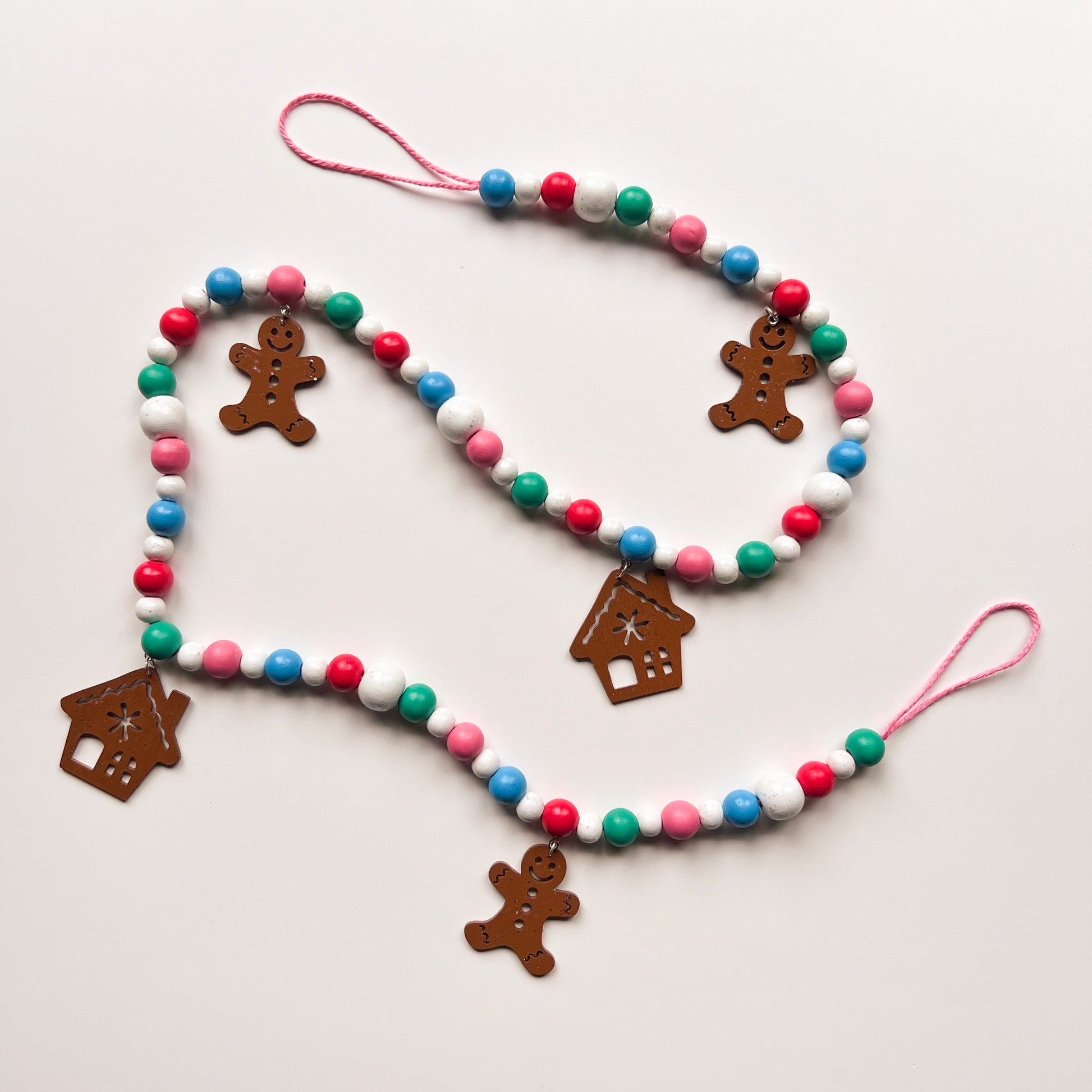 Gingerbread Village Handmade Garland. The garland features three wood gingerbread figures alongside two gingerbread houses. Shapes hang from wood beads. Christmas holiday seasonal decoration or gift.