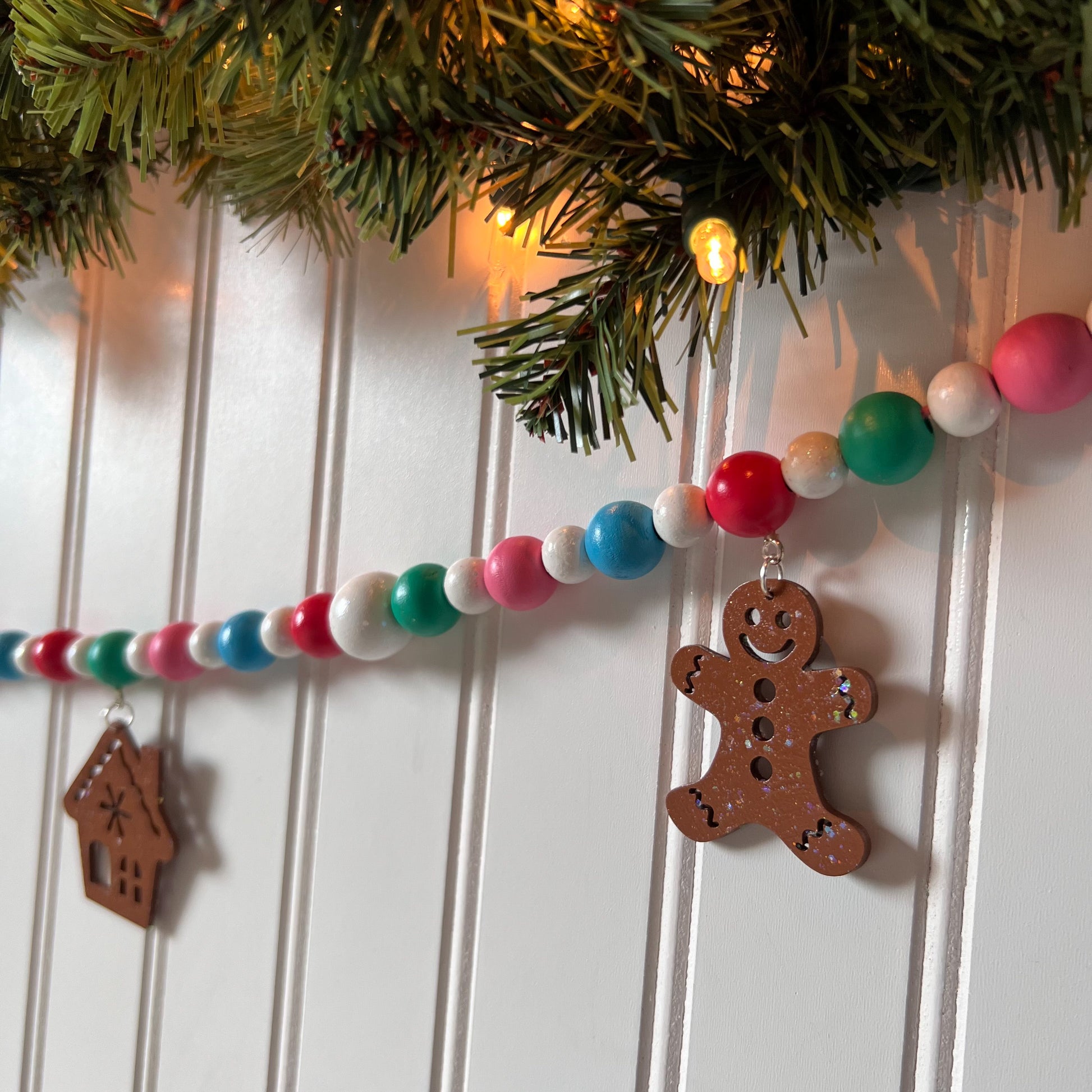 Gingerbread Village Handmade Garland. The garland features three wood gingerbread figures alongside two gingerbread houses. Shapes hang from wood beads. Christmas holiday seasonal decoration or gift.