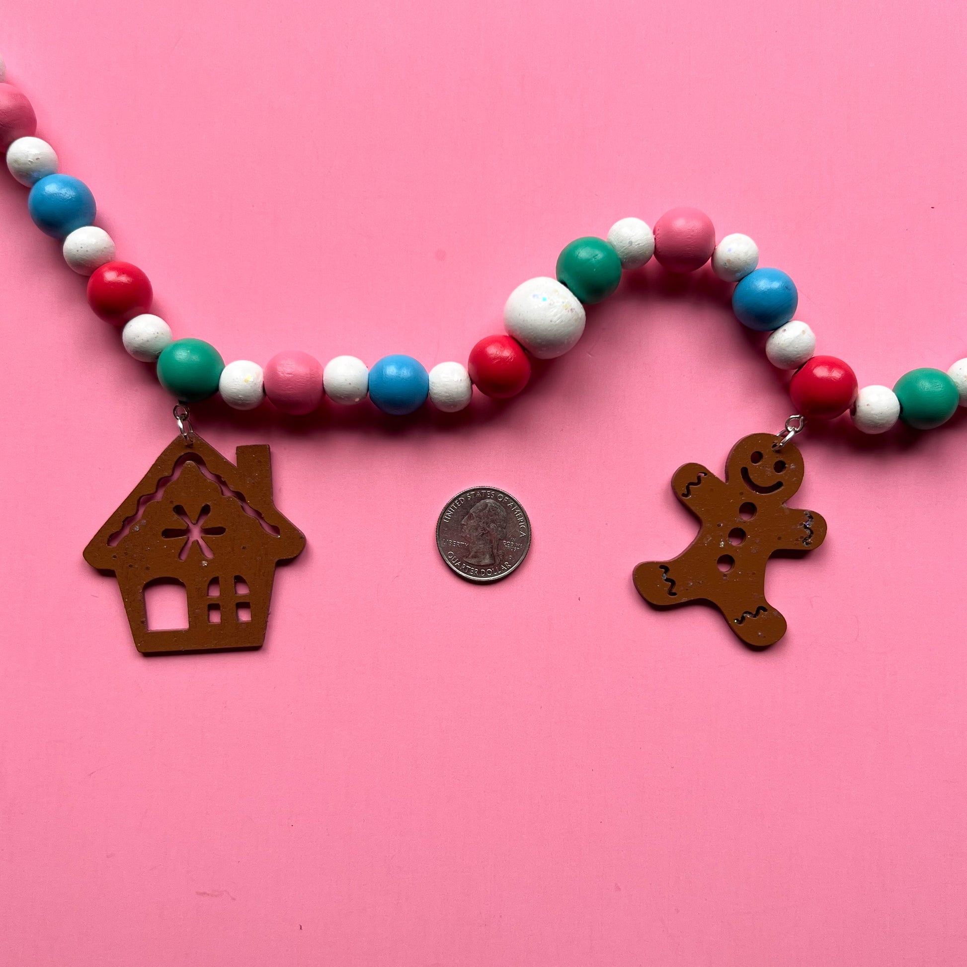 Gingerbread Village Handmade Garland. The garland features three wood gingerbread figures alongside two gingerbread houses. Shapes hang from wood beads. Christmas holiday seasonal decoration or gift.