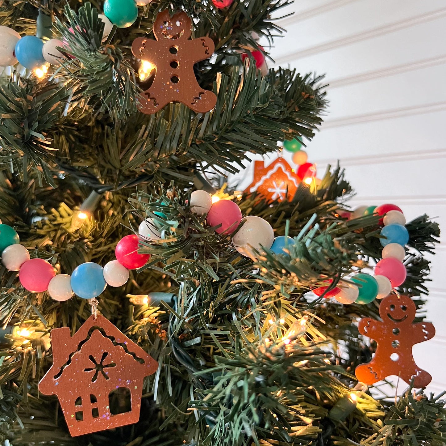 Gingerbread Village Handmade Garland. The garland features three wood gingerbread figures alongside two gingerbread houses. Shapes hang from wood beads. Christmas holiday seasonal decoration or gift.
