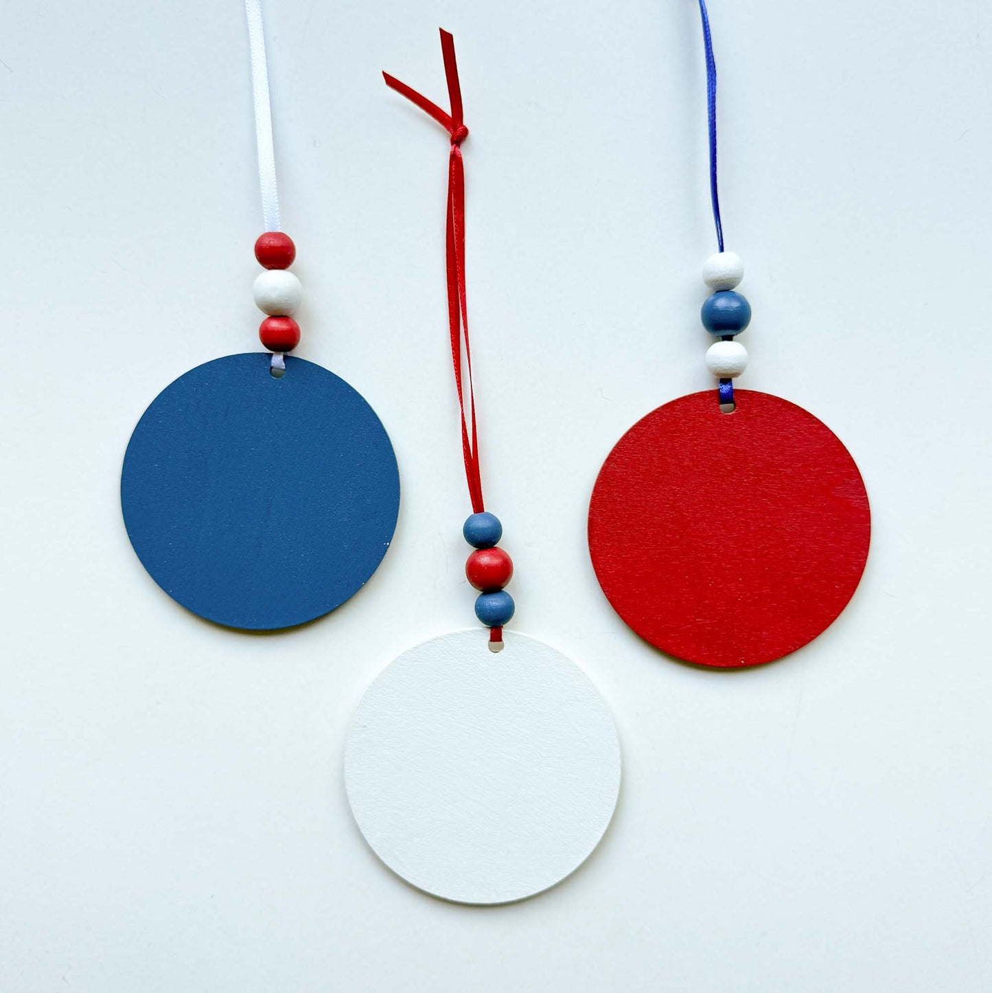 Back of set of 3 patriotic themed ornaments painted in colors of red, white, and blue. The front of the ornaments feature Uncle Sam's hat, "4th of July" cutout, and a firecracker. Ornaments hang from a ribbon with three painted wood beads.