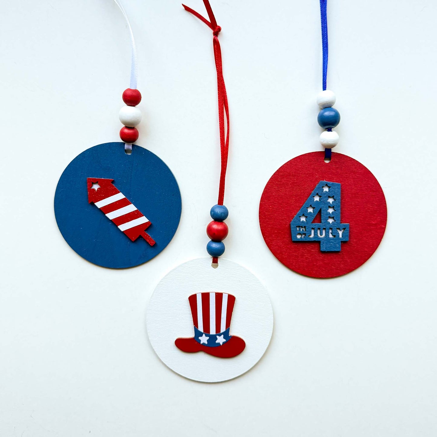 Set of 3 patriotic themed ornaments painted in colors of red, white, and blue. Ornaments feature Uncle Sam's hat, "4th of July" cutout, and a firecracker. Ornaments hang from a ribbon with three painted wood beads.
