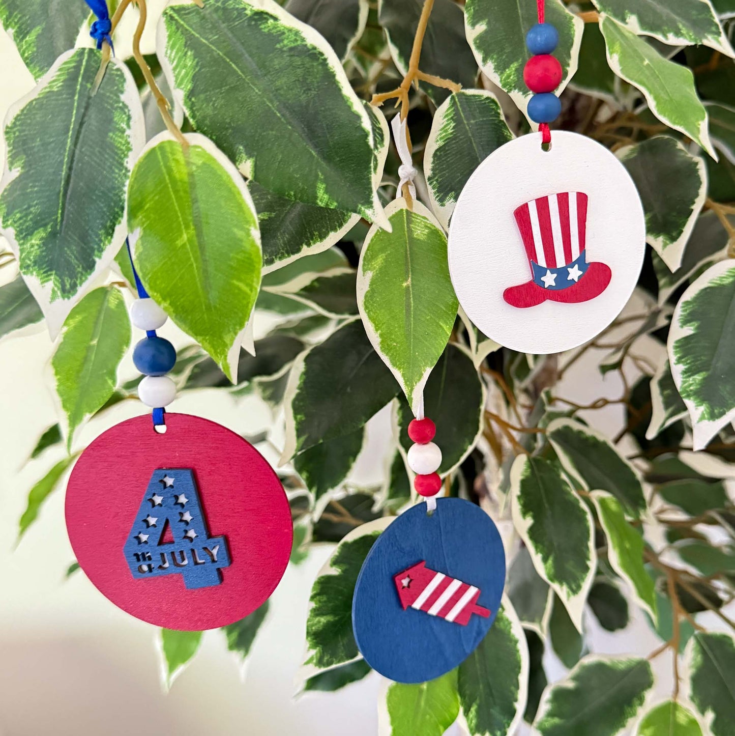 Set of 3 patriotic themed ornaments painted in colors of red, white, and blue. Ornaments feature Uncle Sam's hat, "4th of July" cutout, and a firecracker. Ornaments hang from a ribbon with three painted wood beads.