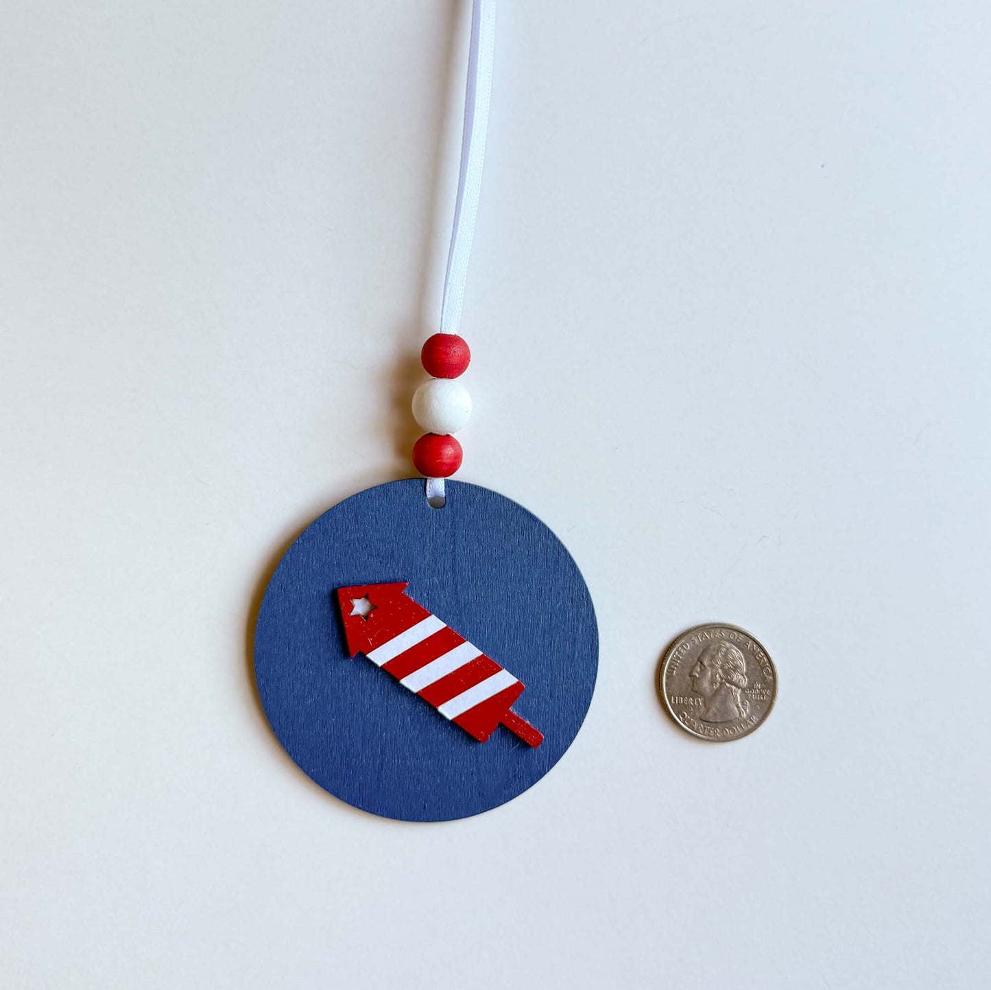 Firecracker 4th of July themed ornament featuring a flat wood shape painted blue, featuring a red and white firecracker. Ornament hangs from a ribbon with three painted wood beads.
