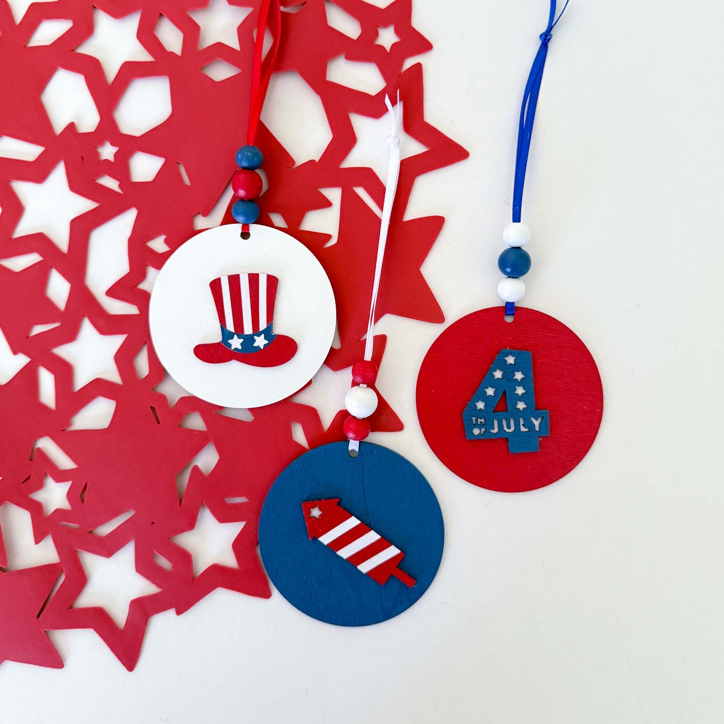 Set of 3 patriotic themed ornaments painted in colors of red, white, and blue. Ornaments feature Uncle Sam's hat, "4th of July" cutout, and a firecracker. Ornaments hang from a ribbon with three painted wood beads.
