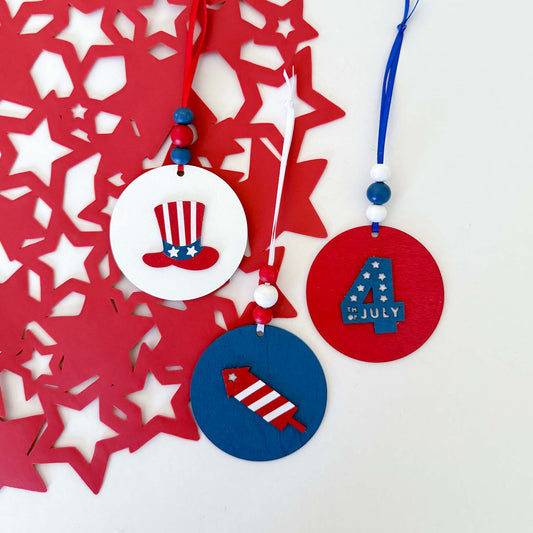 Set of 3 patriotic themed ornaments painted in colors of red, white, and blue. Ornaments feature Uncle Sam's hat,  "4th of July" cutout, and a firecracker. Ornaments hang from a ribbon with three painted wood beads.