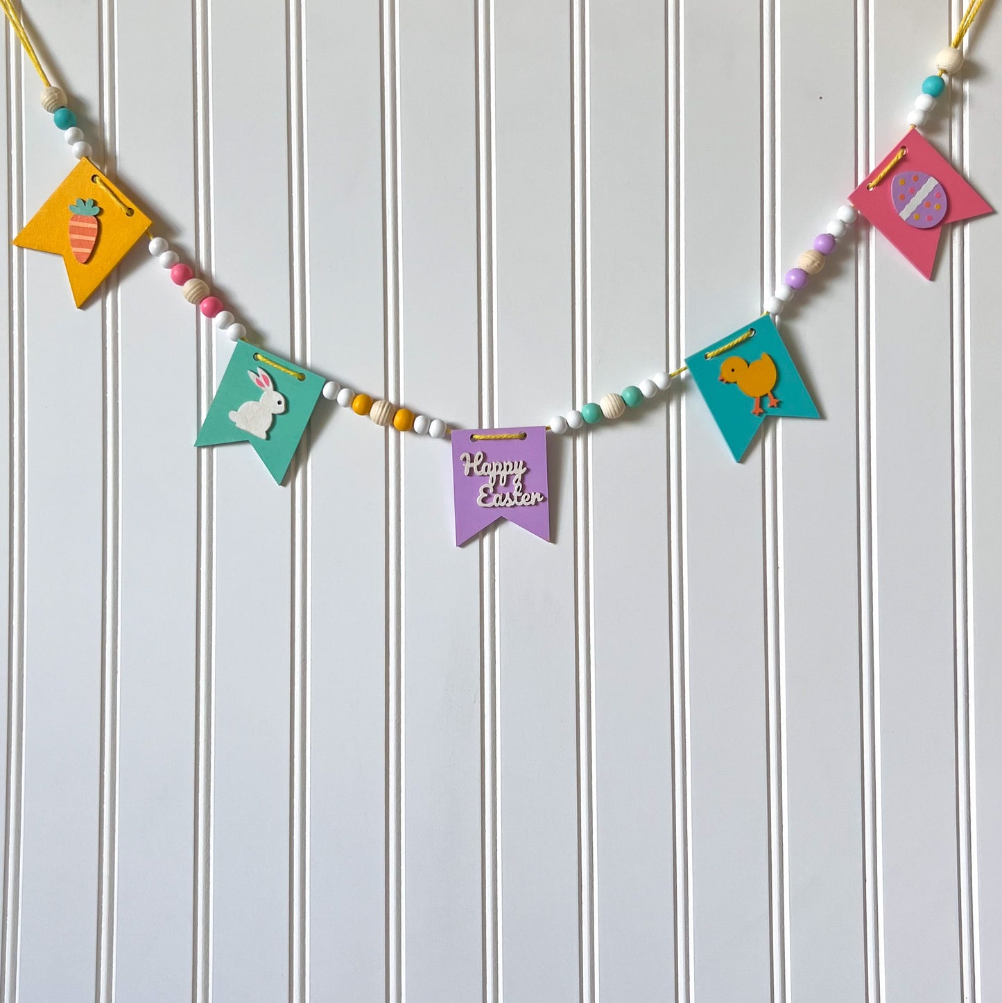 Image shows handmade garland with wood pennants featuring Easter shapes - a carrot, a bunny, a chick, an egg, and a "Happy Easter" wood cutout. Pennants are strung with natural and painted beads; includes 3-inch loops on either end.