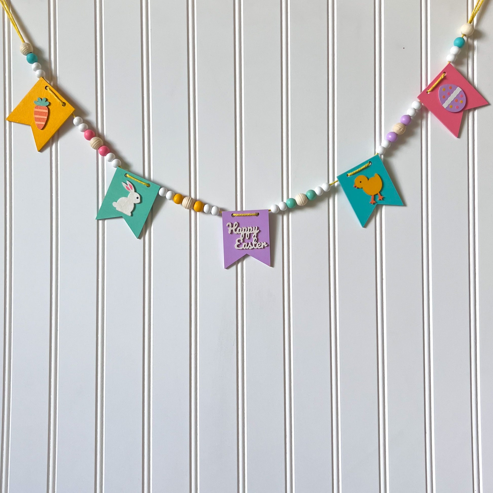 Image shows handmade garland with wood pennants featuring Easter shapes - a carrot, a bunny, a chick, an egg, and a "Happy Easter" wood cutout. Pennants are strung with natural and painted beads; includes 3-inch loops on either end.