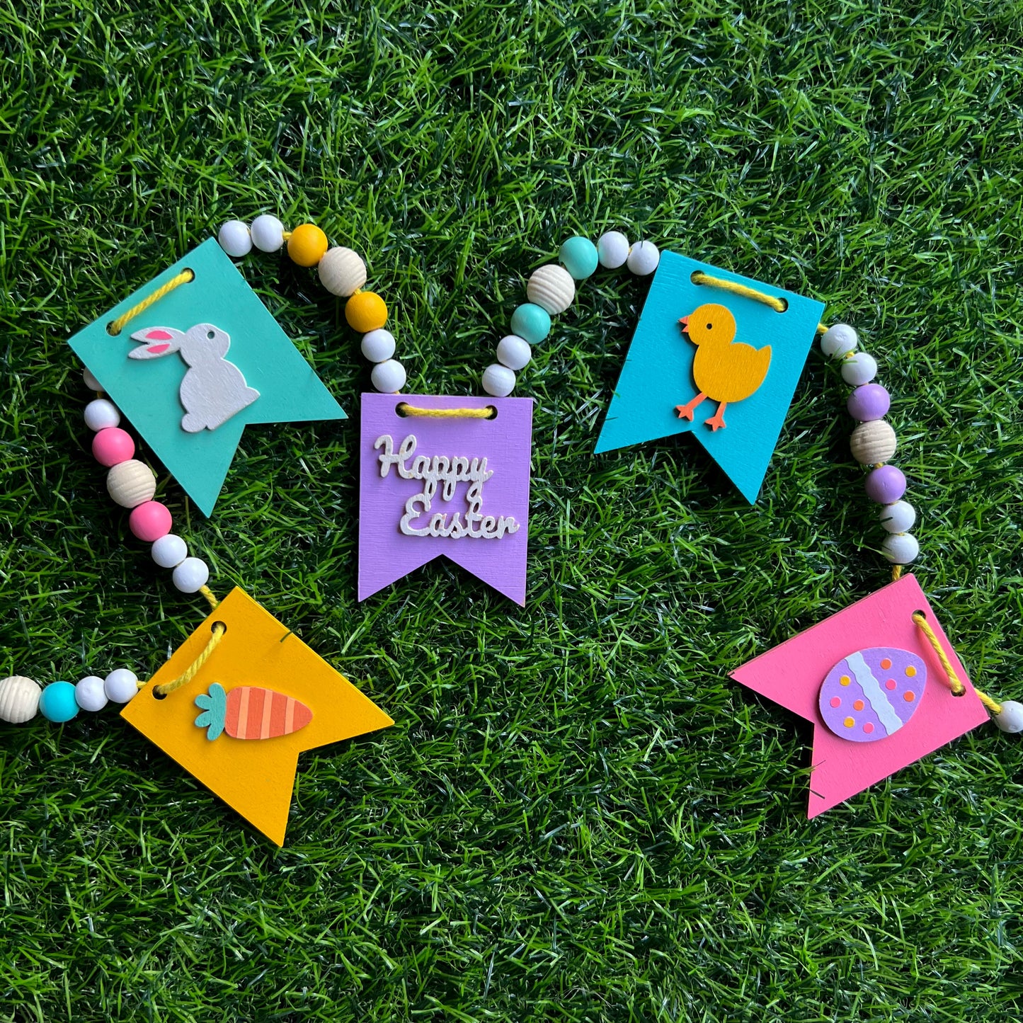 Image shows handmade garland with wood pennants featuring Easter shapes - a carrot, a bunny, a chick, an egg, and a "Happy Easter" wood cutout. Pennants are strung with natural and painted beads; includes 3-inch loops on either end.