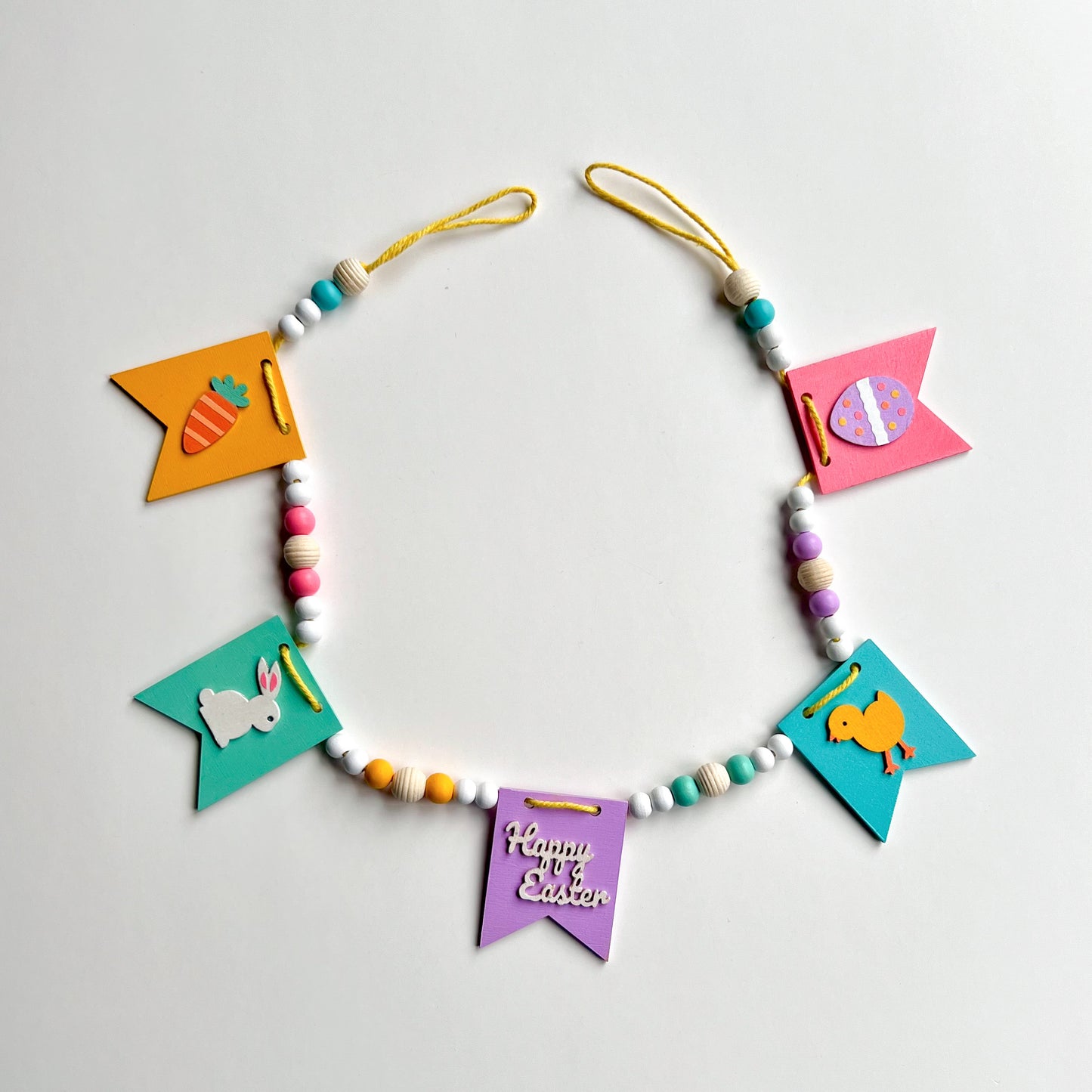 Image shows handmade garland with wood pennants featuring Easter shapes - a carrot, a bunny, a chick, an egg, and a "Happy Easter" wood cutout. Pennants are strung with natural and painted beads; includes 3-inch loops on either end.