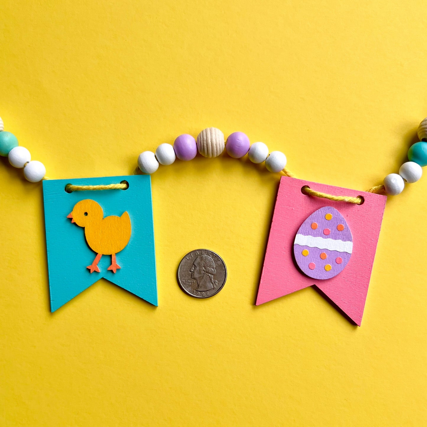Closeup of image showing handmade garland with wood pennants featuring Easter shapes - a chick and an egg. Pennants are strung with natural and painted beads; includes 3-inch loops on either end.