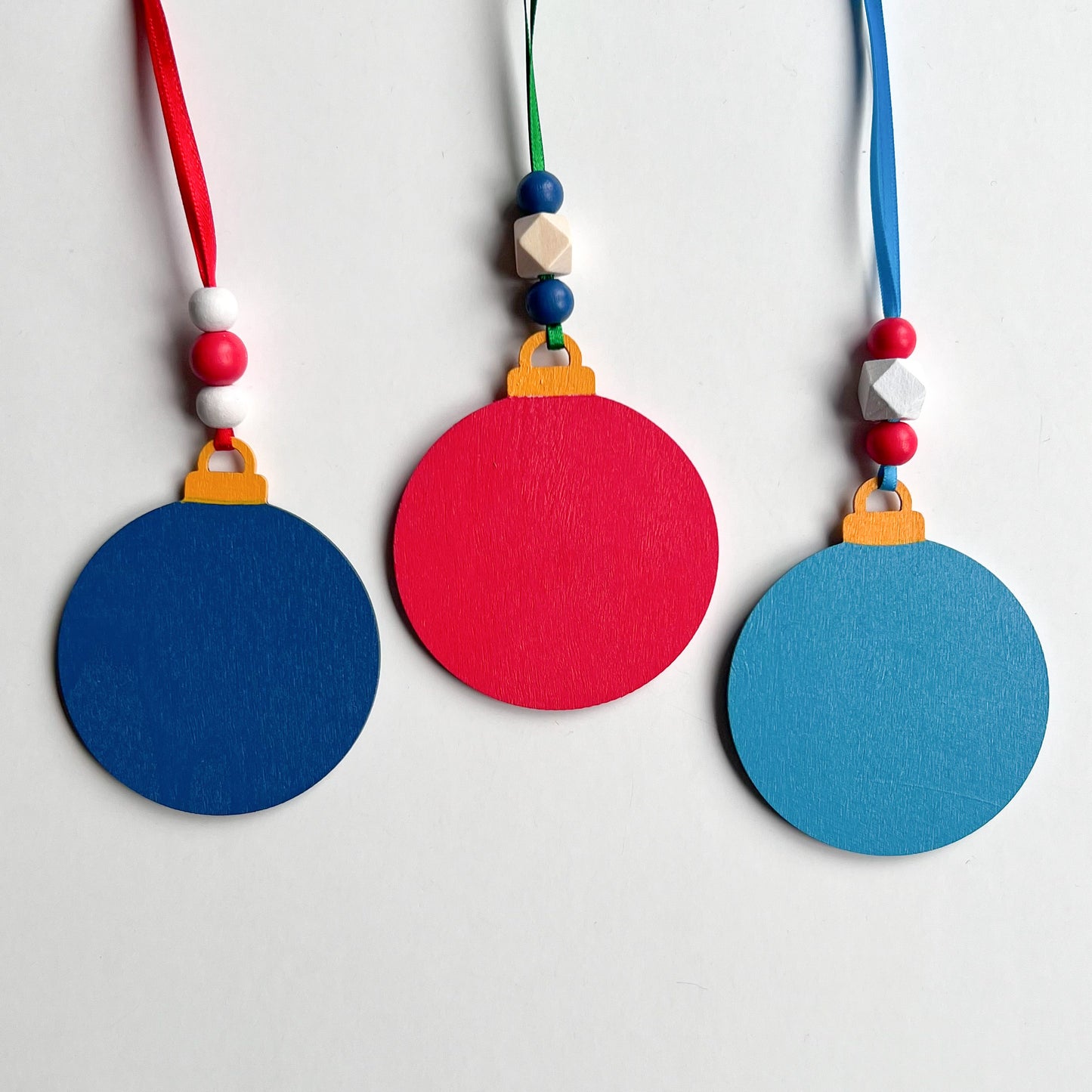 Image shows the back of a set of 3 flat wood handmade ornaments. The front of each ornament features a holiday themed wood shape: a snowman, a candy cane, or a truck bringing home the tree. Ornaments hang from ribbon and are decorated with three wood beads in matching colors.