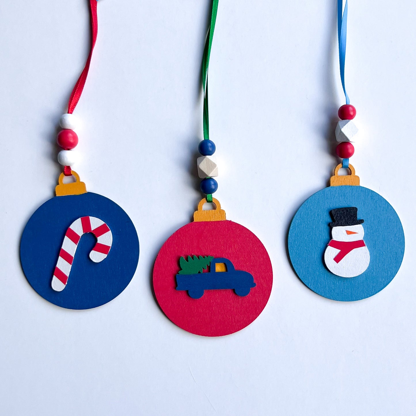 Image shows set of 3 flat wood handmade ornaments. Each ornament features a holiday themed wood shape: a snowman, a candy cane, or a truck bringing home the tree. Ornaments hang from ribbon and are decorated with three wood beads in matching colors.