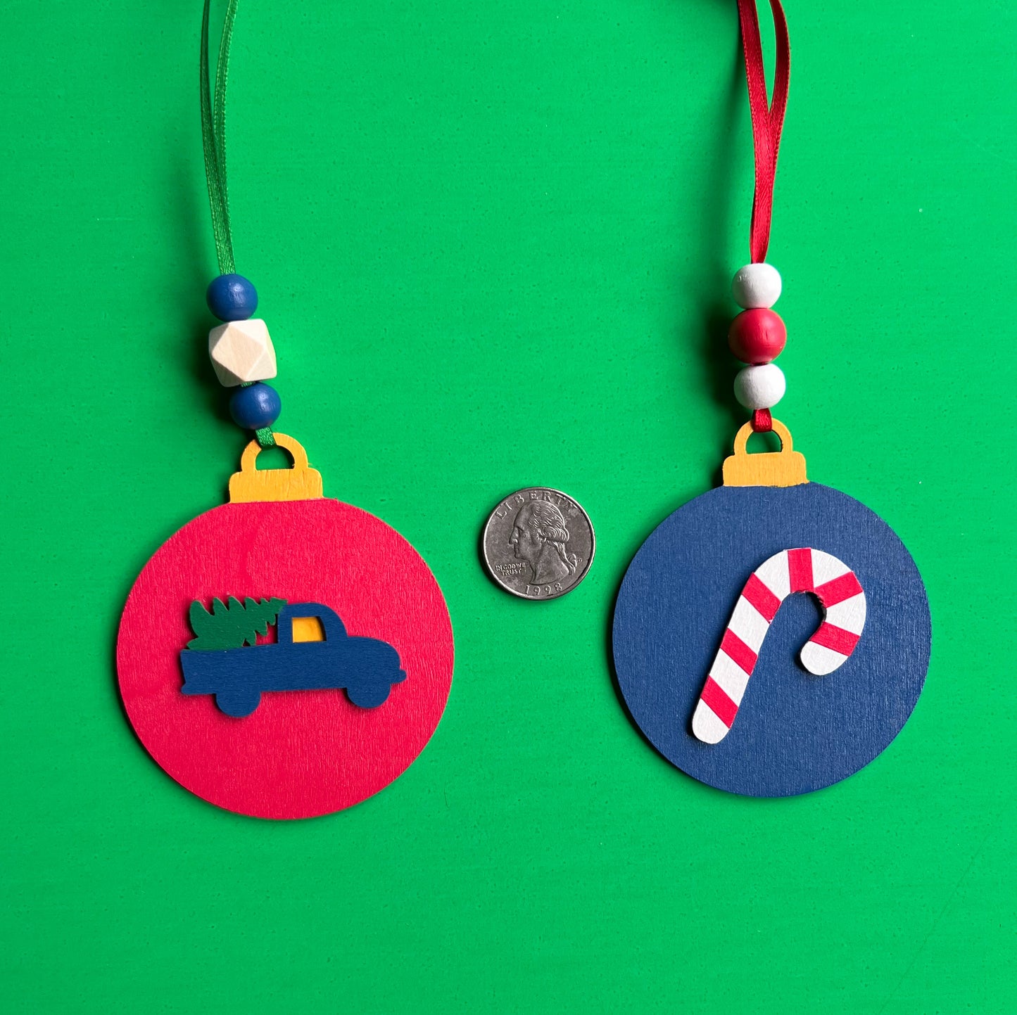 Image shows set of flat woures a holiday theod handmade ornaments. Each ornament featmed wood shape: a candy cane or a truck bringing home the tree. Ornaments hang from ribbon and are decorated with three wood beads in matching colors.