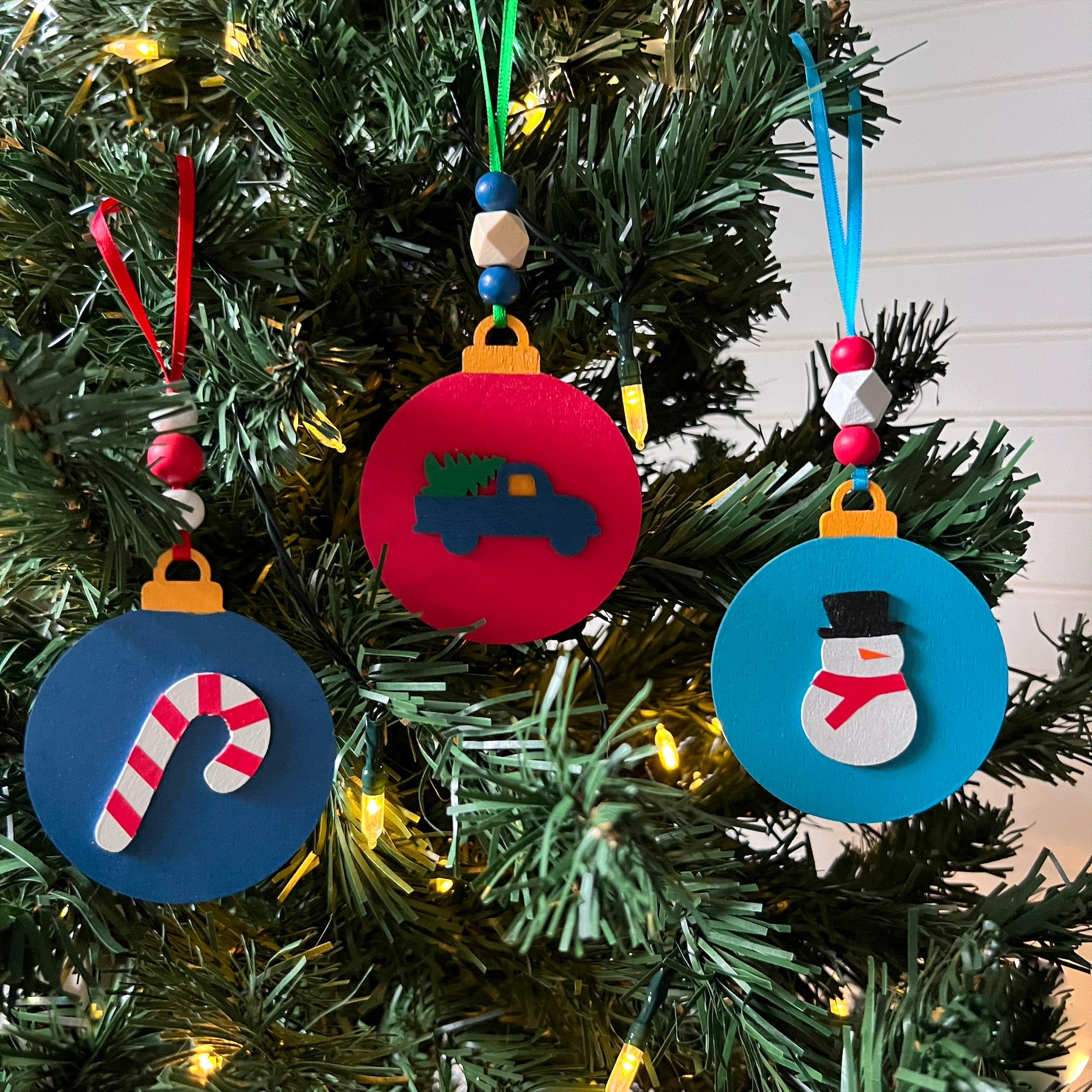 Image shows set of 3 flat wood handmade ornaments. Each ornament features a holiday themed wood shape: a snowman, a candy cane, or a truck bringing home the tree. Ornaments hang from ribbon and are decorated with three wood beads in matching colors.